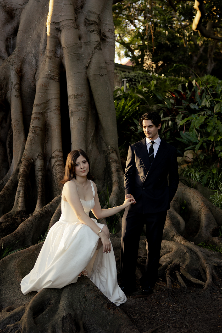 A woman in a white dress and a man in a black suit standing in a forest near a large tree with sprawling roots.