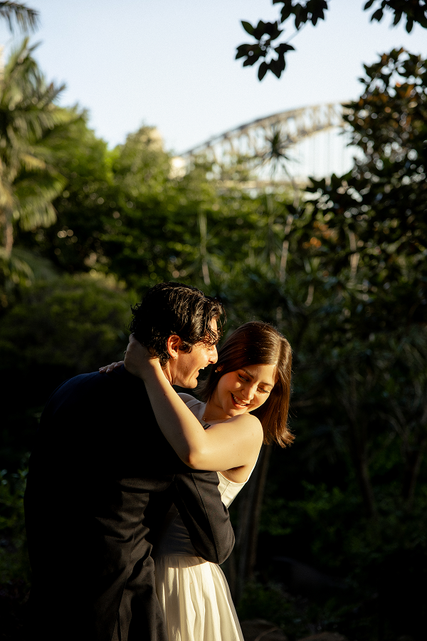 A couple embracing outdoors during daytime, with trees and a bridge in the background.