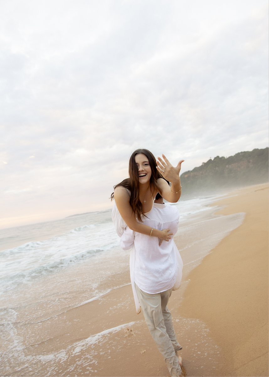 A young woman with long brown hair smiling and waving on a sandy beach, wearing a white top and beige pants, with ocean waves and cloudy sky in the background.