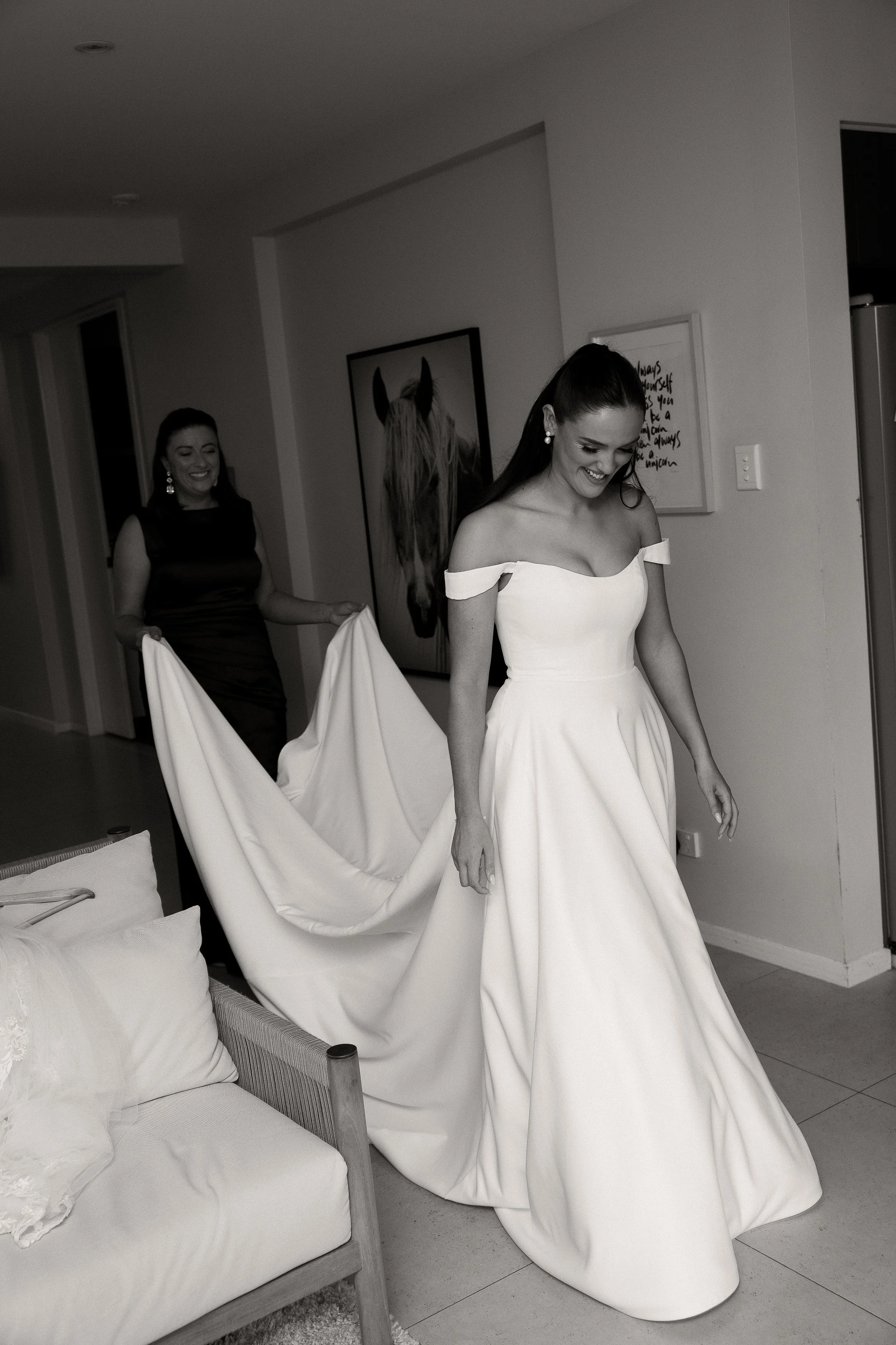 A bride walks with her mother through her house moments after puttingon her wedding dress. Her mother carries the back of her white dress photographed by Haze Photo and Film