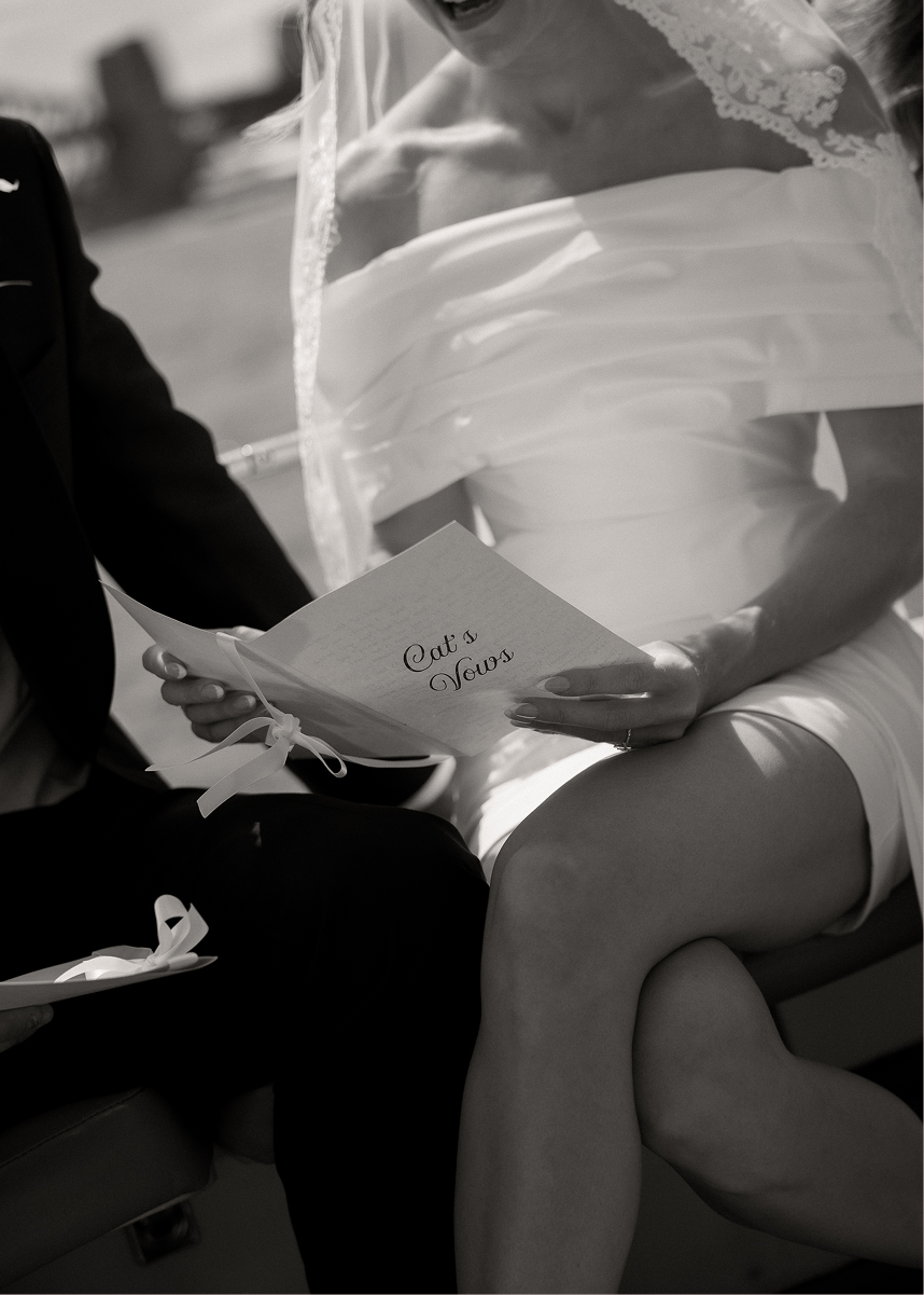 A bride and groom sitting close together, with the bride holding a card that says "Cat's Vows" during a wedding ceremony or reception.