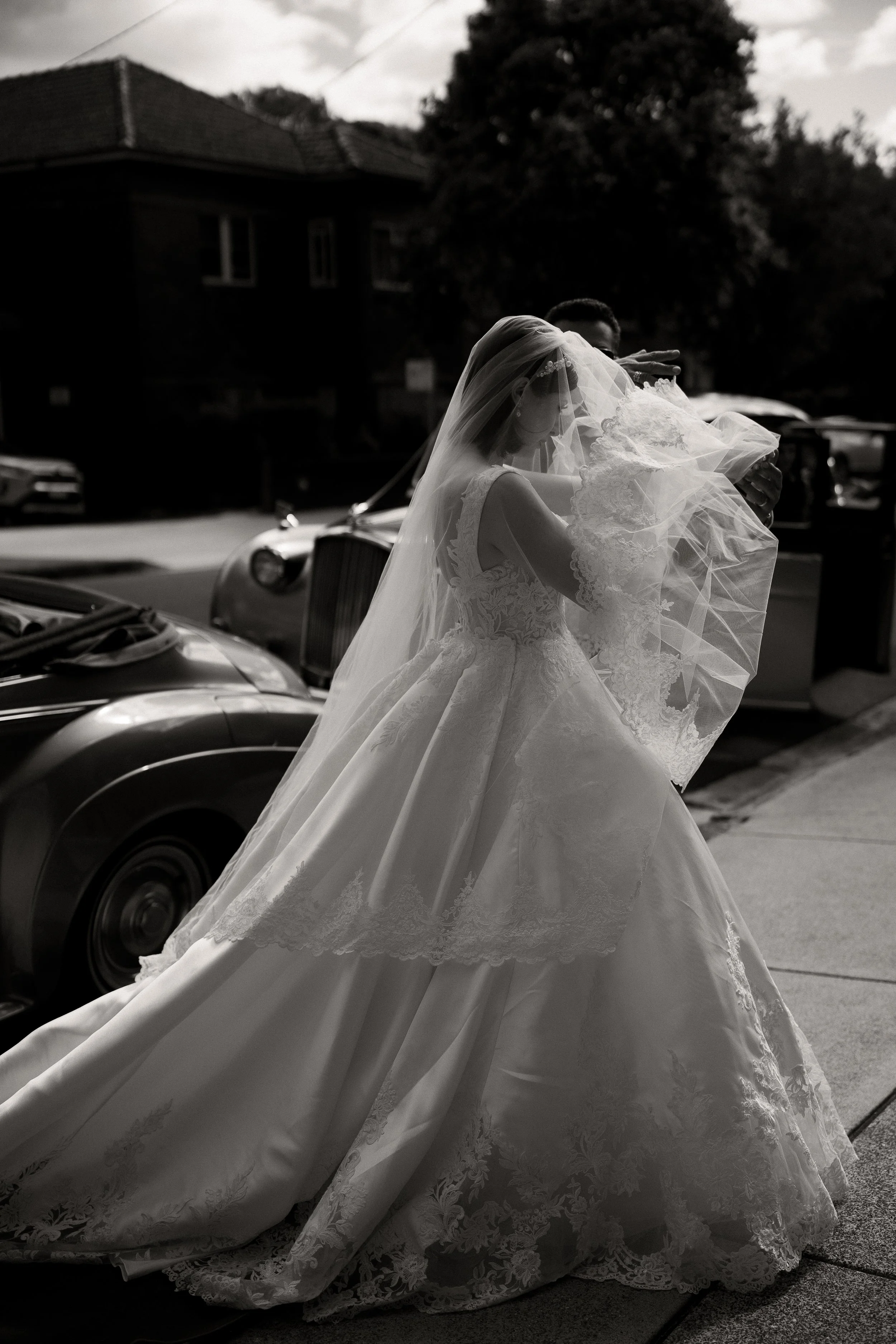 A bride arrives at her ceremony venue and is pulled her viel over her face. She has just come out of a Roll Royce and her father stands behind her getting ready to walk down the aisle, photographed by Haze Photo and Film