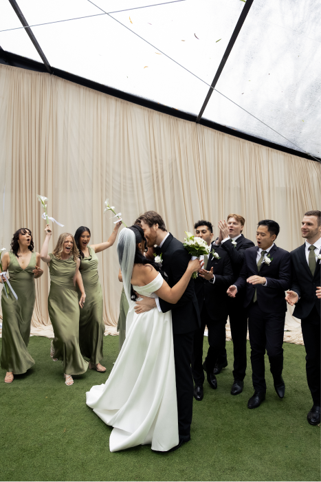 Bridal couple kissing, surrounded by wedding party celebrating indoors with beige curtains and a green carpet.