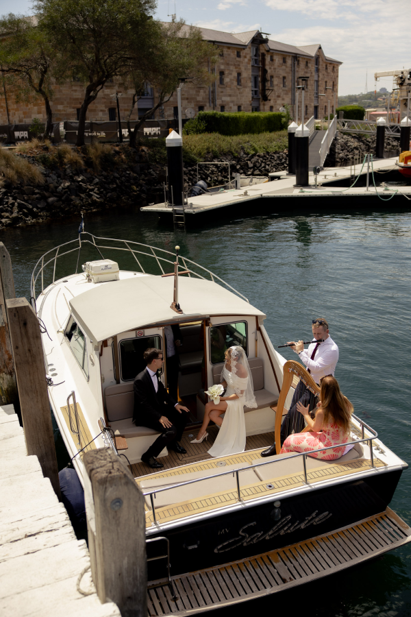 A couple in wedding attire on a boat with a woman playing a harp and a man playing a flute, near a harbor with buildings and trees in the background.