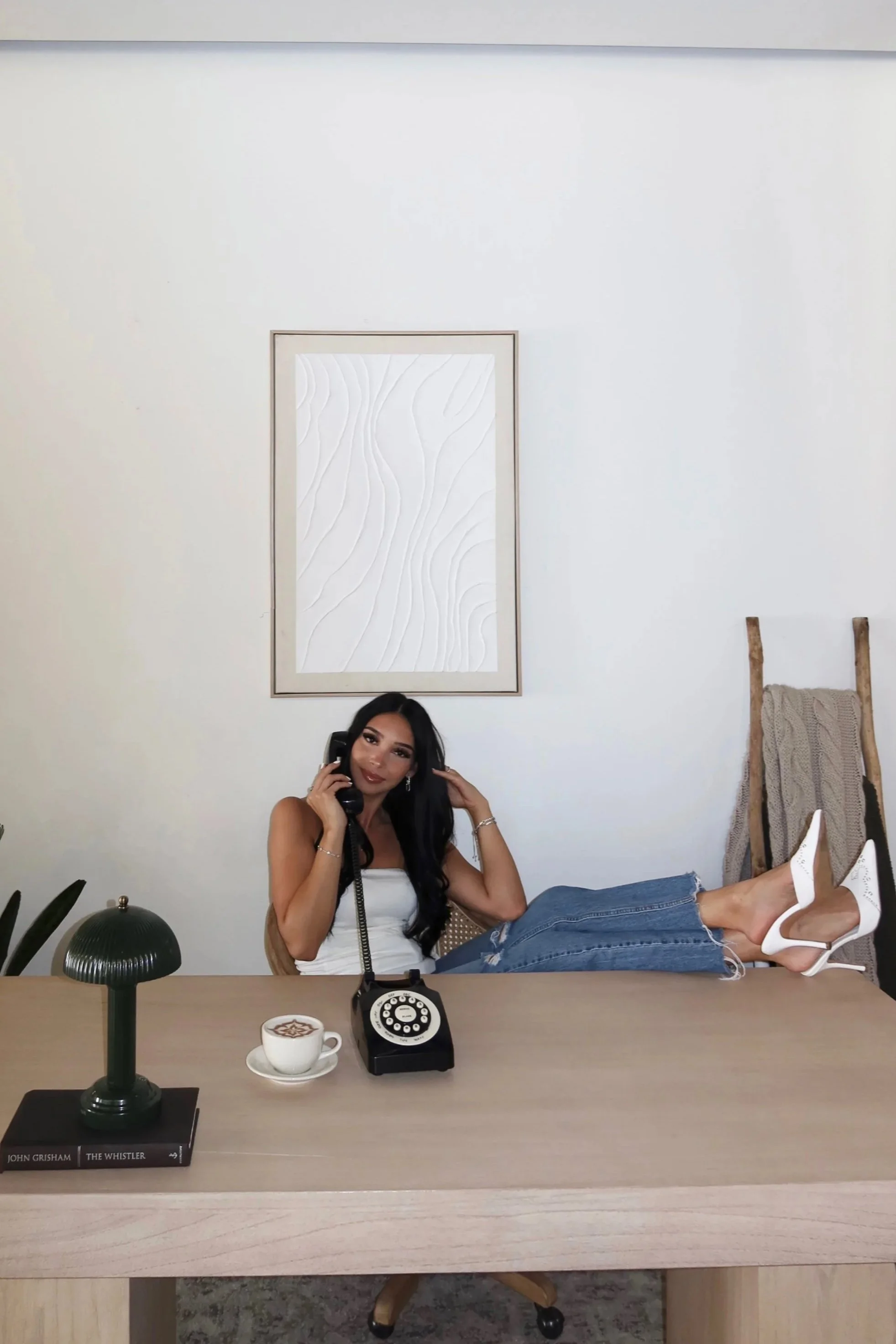 A woman with long black hair sitting at a desk, talking on a vintage black rotary phone. She is wearing a white sleeveless top, ripped jeans, and white high heels. There is a cup of coffee and a green table lamp on the desk. The background features a white wall with a framed abstract art piece and a wooden ladder with a knitted blanket draped over it.