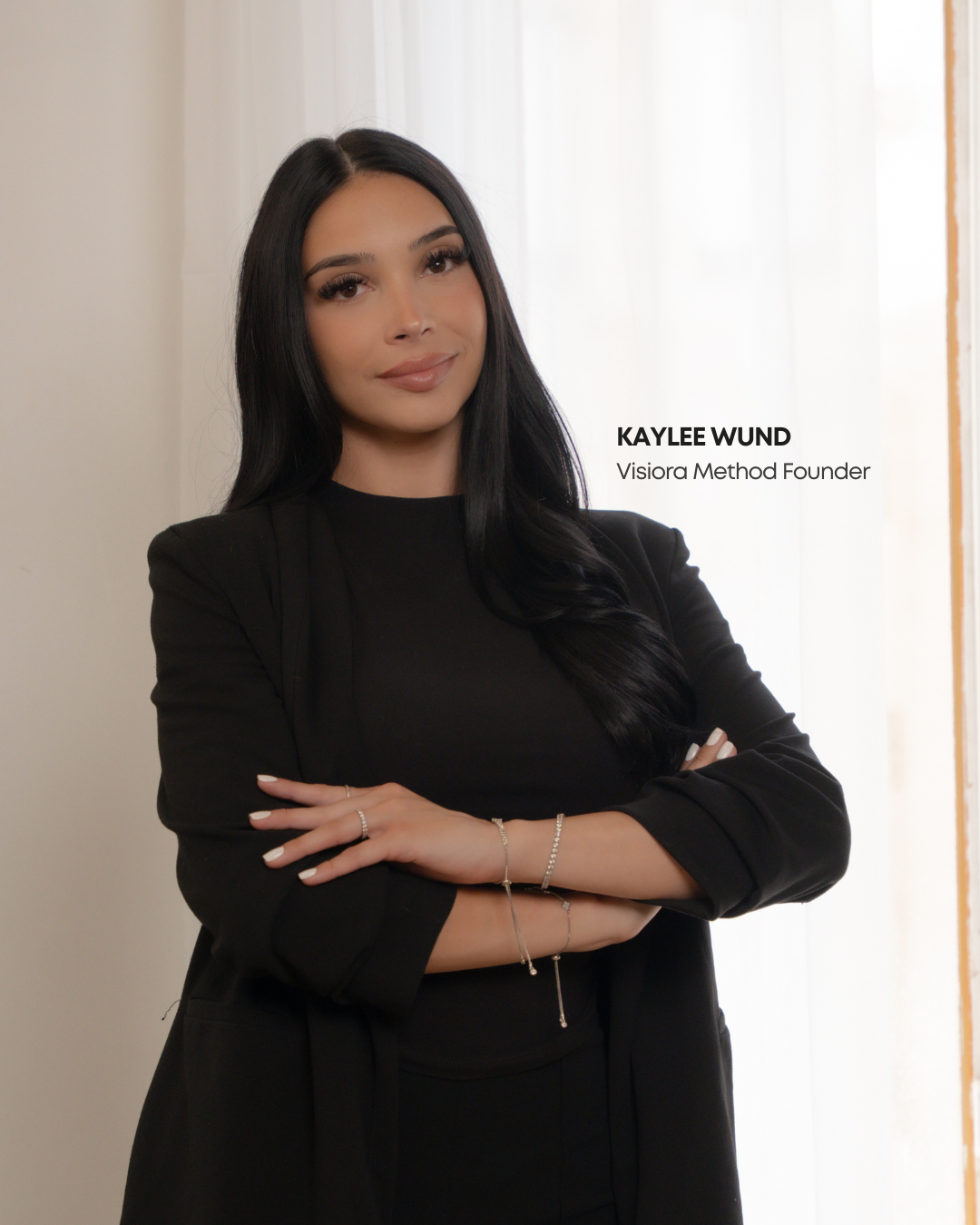 A woman with long dark hair wearing a black blazer and top standing with arms crossed in front of a white curtain. Text overlay reads "KAYLEE WUND, Visiora Method Founder."