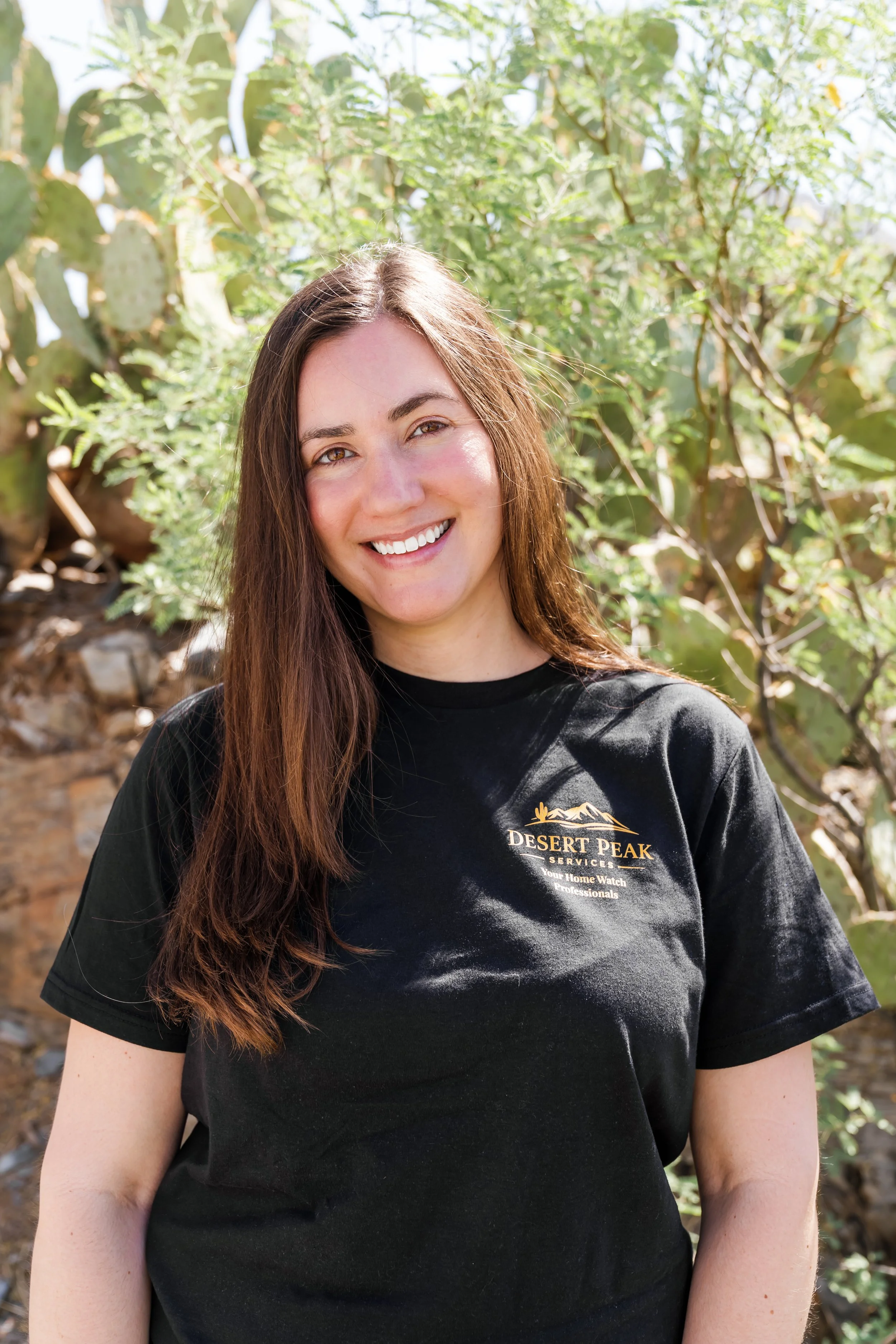 A smiling woman with long brown hair wearing a black Desert Peak Services T-shirt standing outdoors in front of cactus plants.