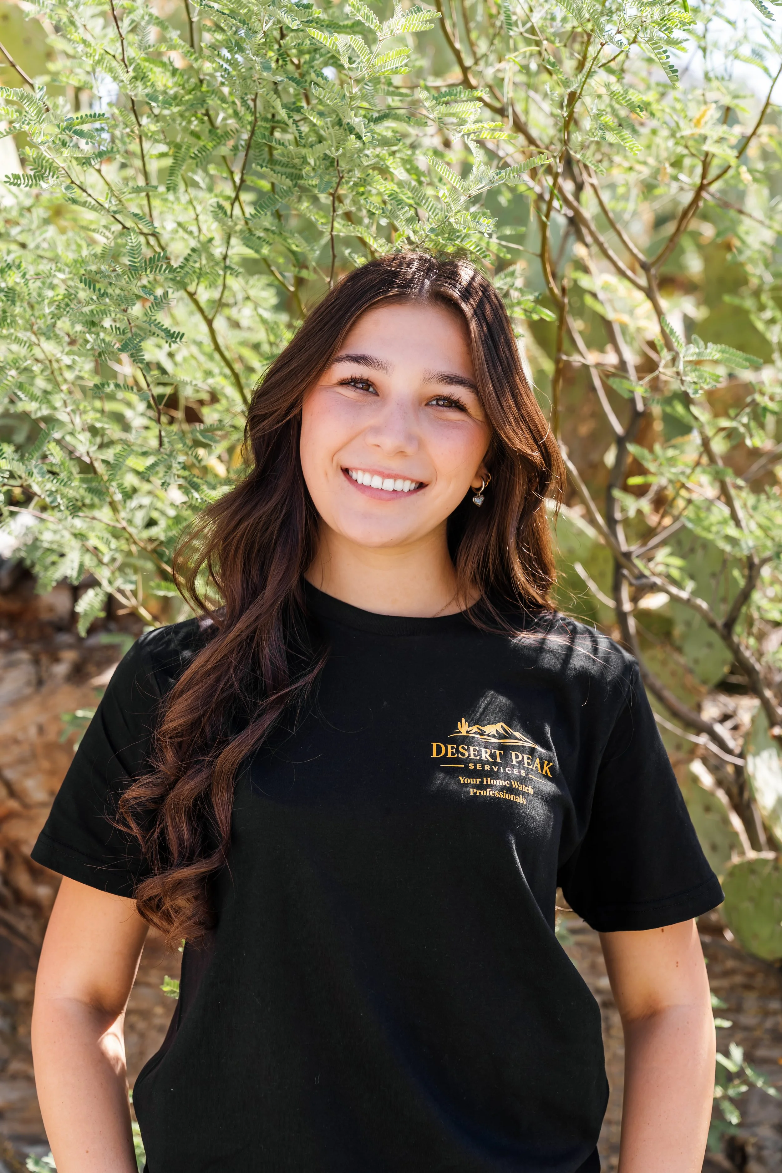 A young woman with long brown hair and earrings smiling outdoors in front of green leafy plants. She is wearing a black t-shirt with a desert landscape logo and the text 'Desert Peak Services, Your Home Watch Professionals'.
