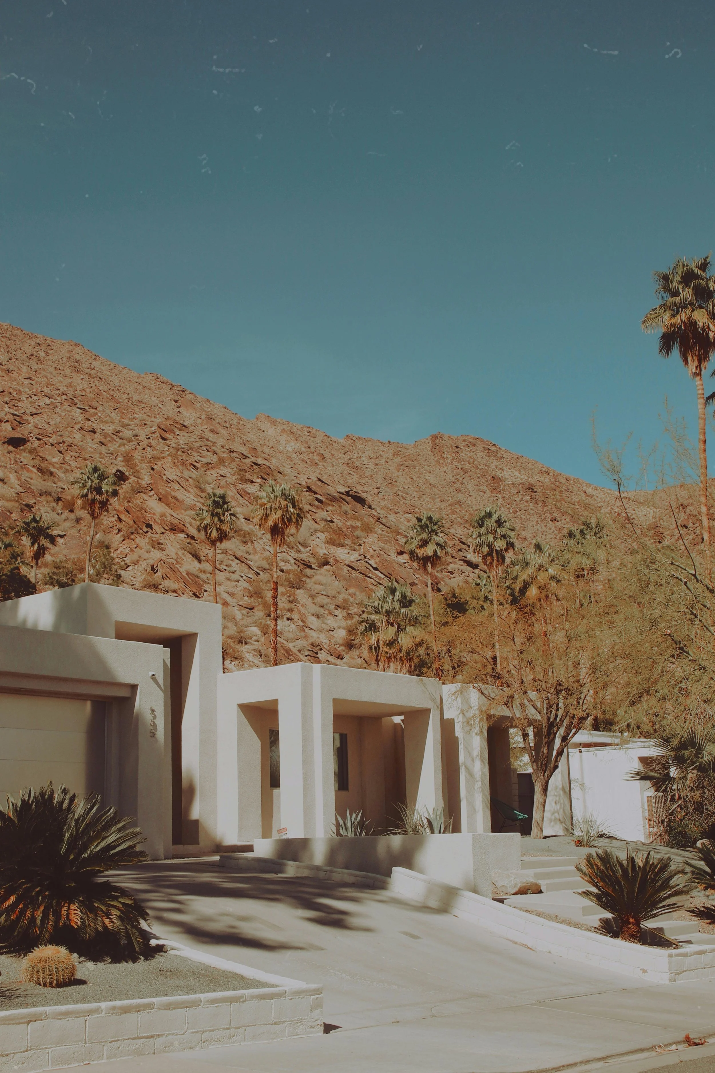 Modern white residential buildings with flat roofs and rectangular shapes, set against a hillside with sparse vegetation and palm trees, under a clear blue sky.