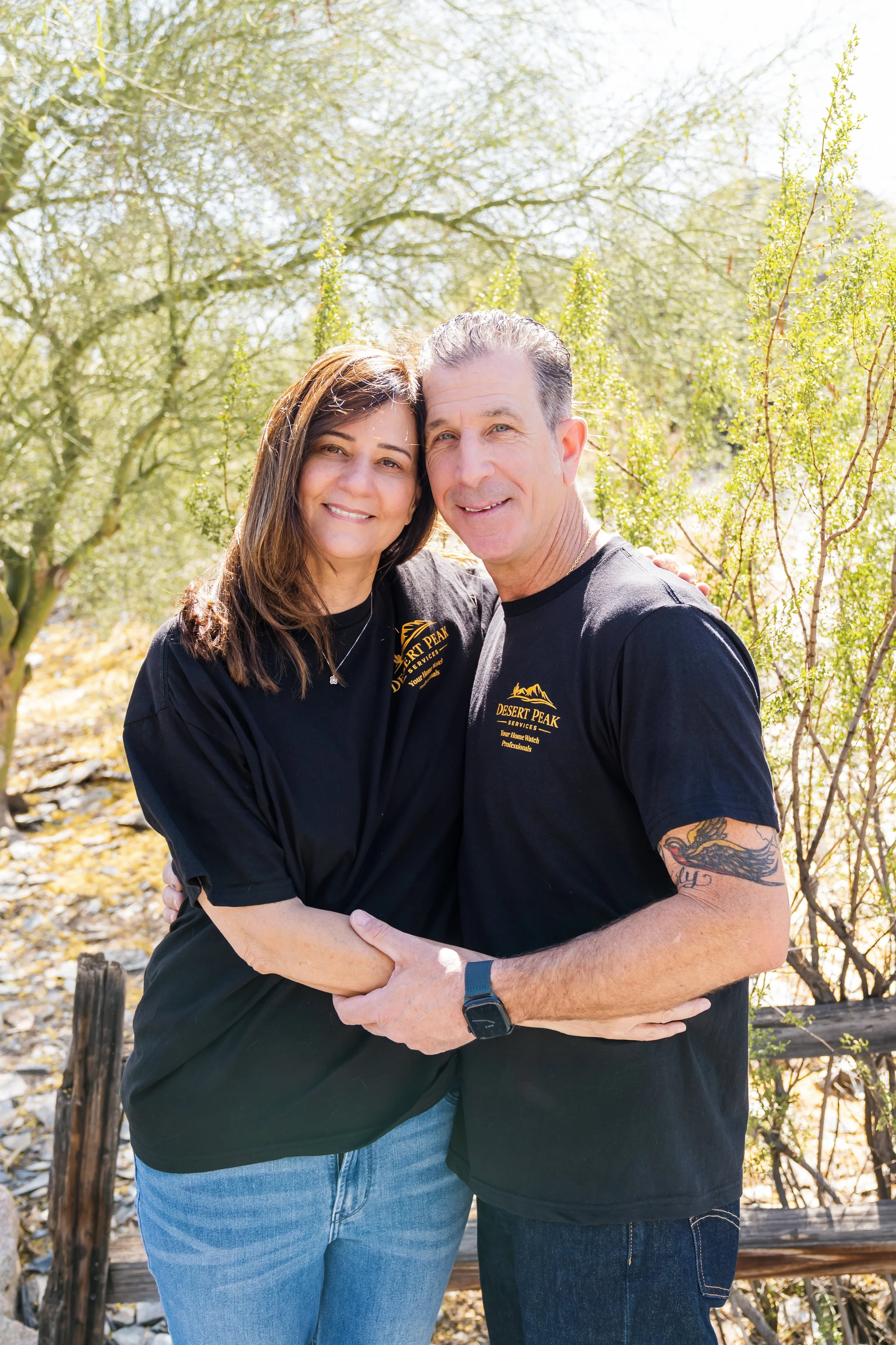 A smiling middle-aged couple hugging outdoors in a sunny park with trees and foliage in the background. Both are wearing black shirts with yellow logos.