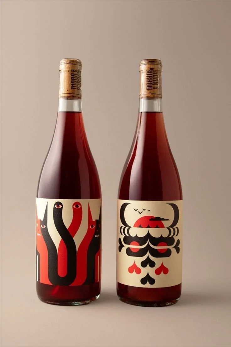 Two bottles of red wine with artistic labels, one featuring a black and red snake design and the other with a stylized blood drip and black hearts.