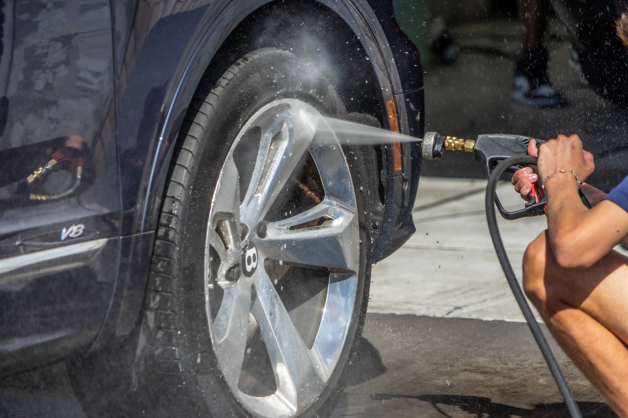 Person washing a black luxury car, focusing on the front wheel, with a pressure washer.