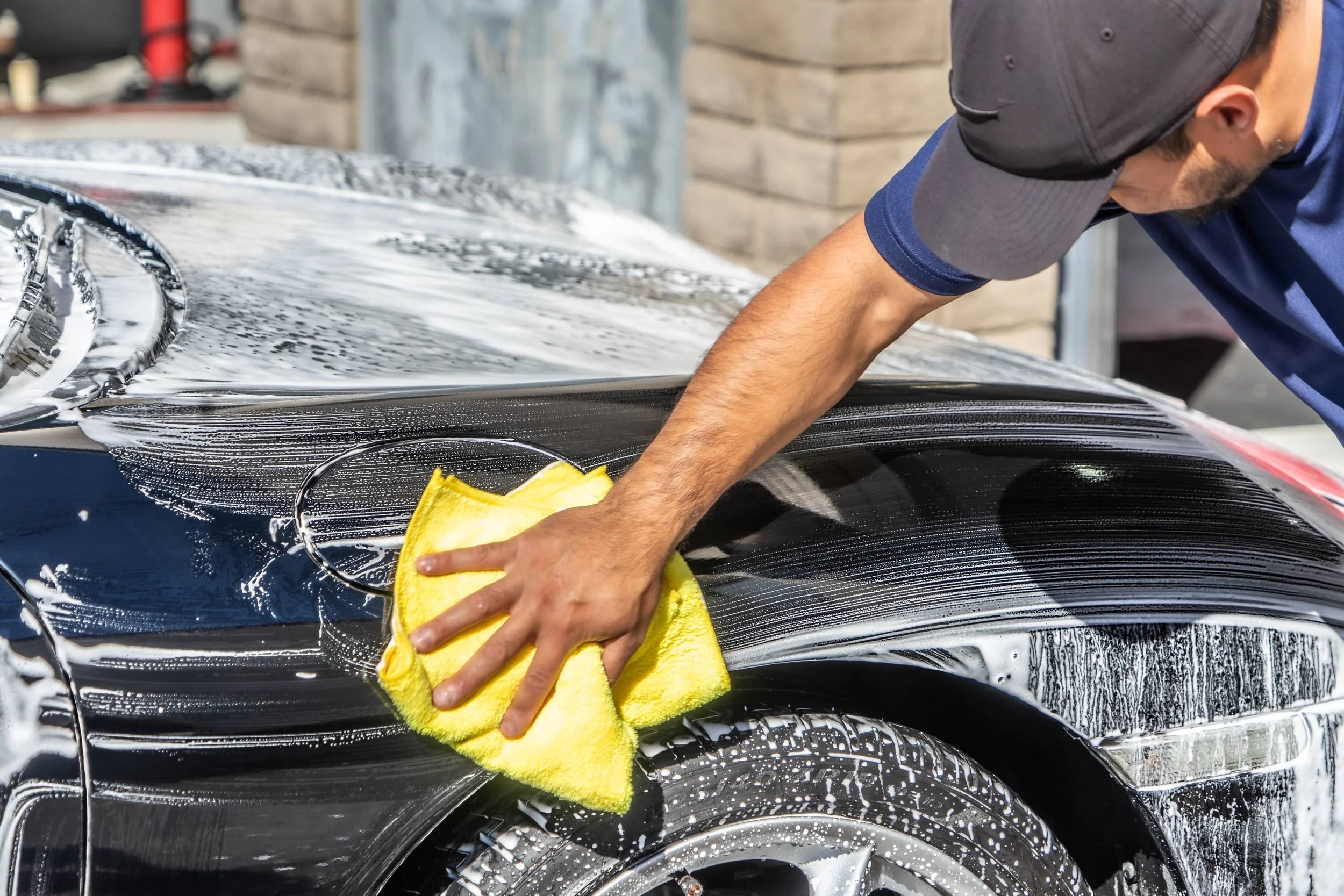 A man washing a black car with a yellow sponge, creating soap suds on the vehicle's surface.