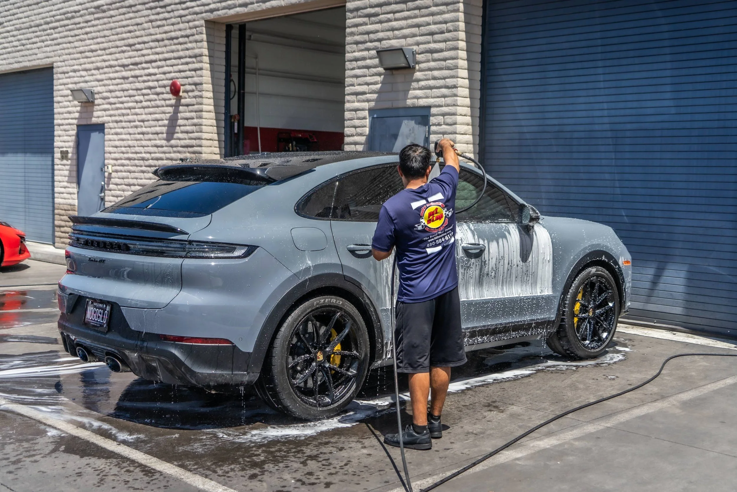 Man washing a silver Porsche SUV with a hose outside a building.