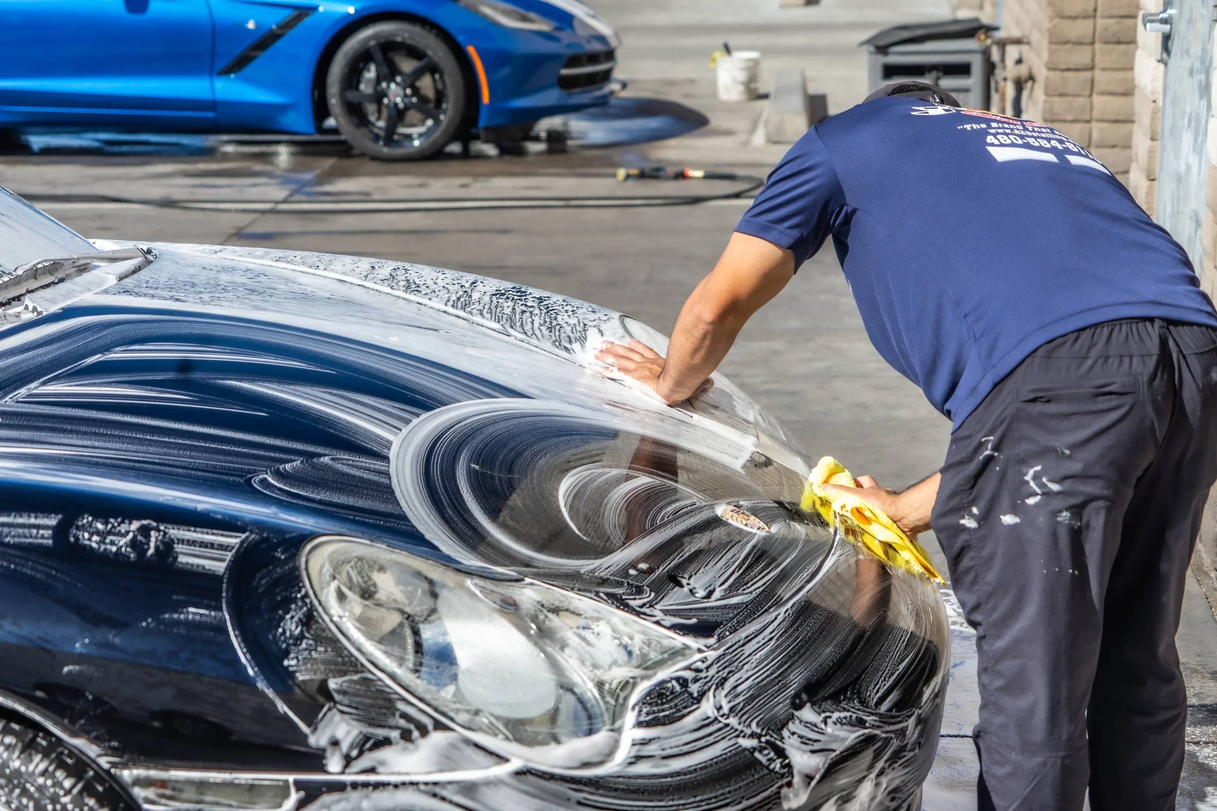 Man washing a black car with soap and a yellow sponge in a driveway, with a blue sports car in the background.