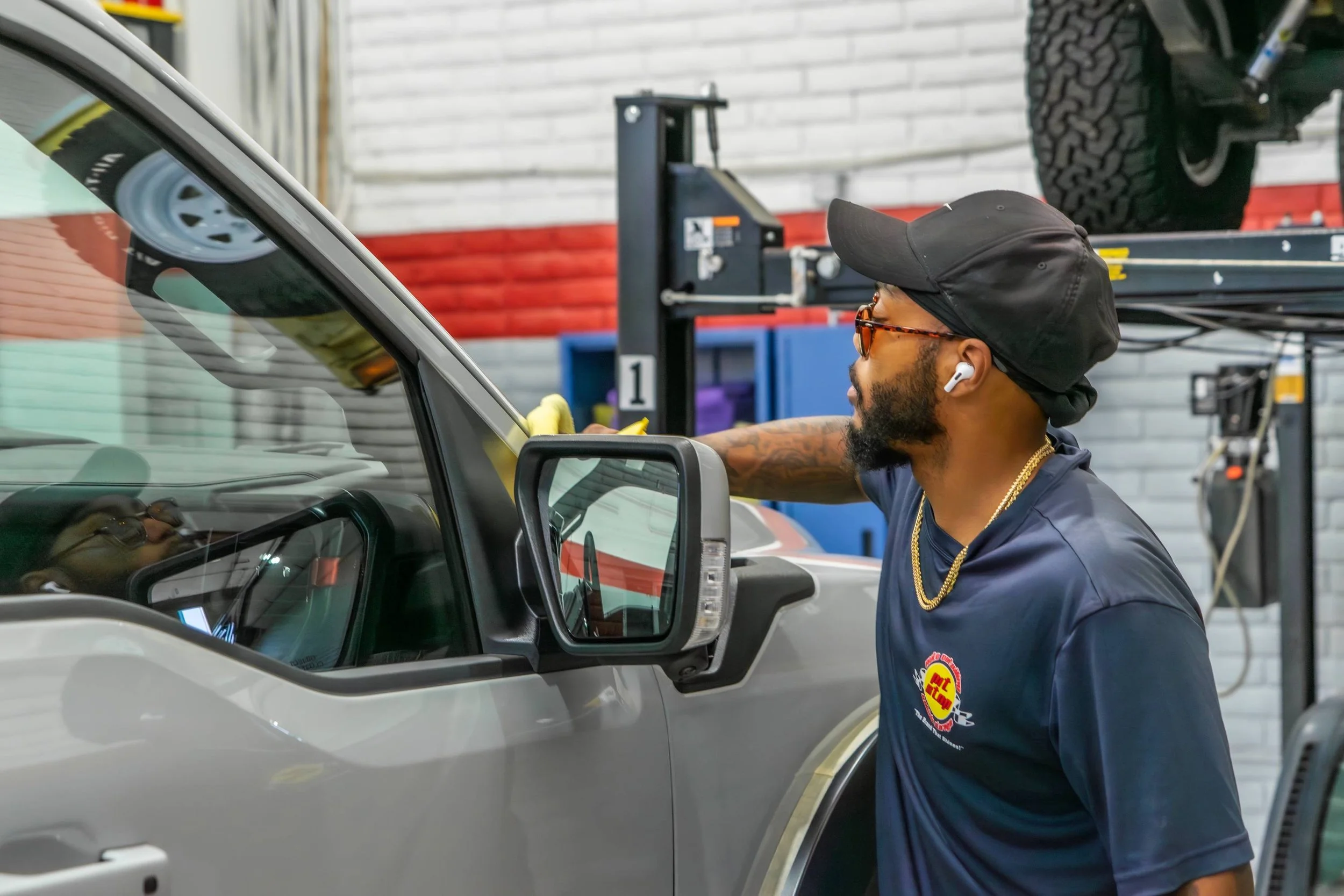 A man working on a silver vehicle in a garage, wearing a black cap, sunglasses, a navy blue t-shirt, and wireless earbuds.
