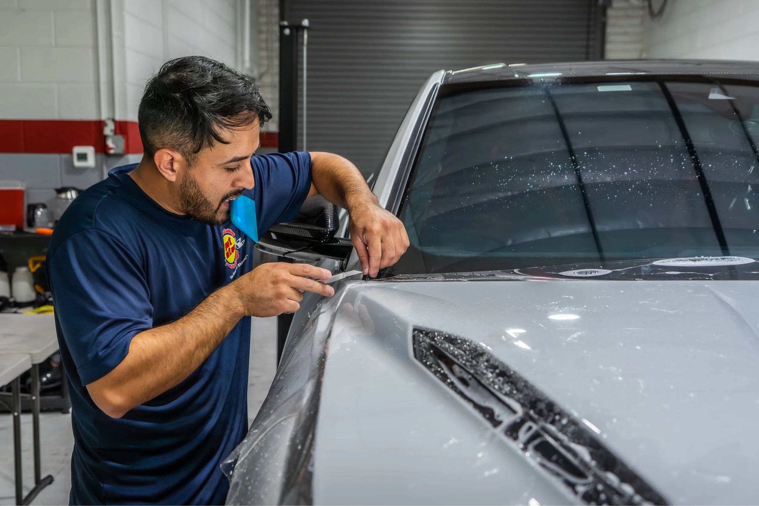 A man in a blue shirt installs a clear protective film on the hood of a silver sports car in a garage.