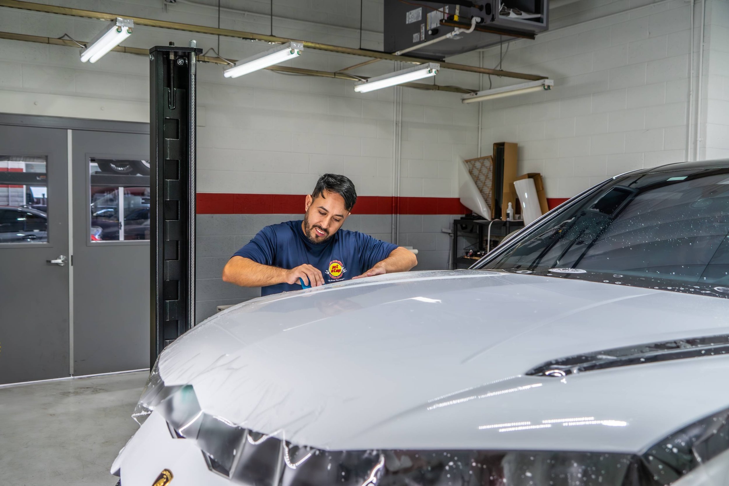 A man polishing a silver sports car inside a garage. The man is wearing a blue t-shirt and appears to be cleaning or polishing the car's hood with a cloth. The garage has fluorescent lighting and shelves with various items in the background.
