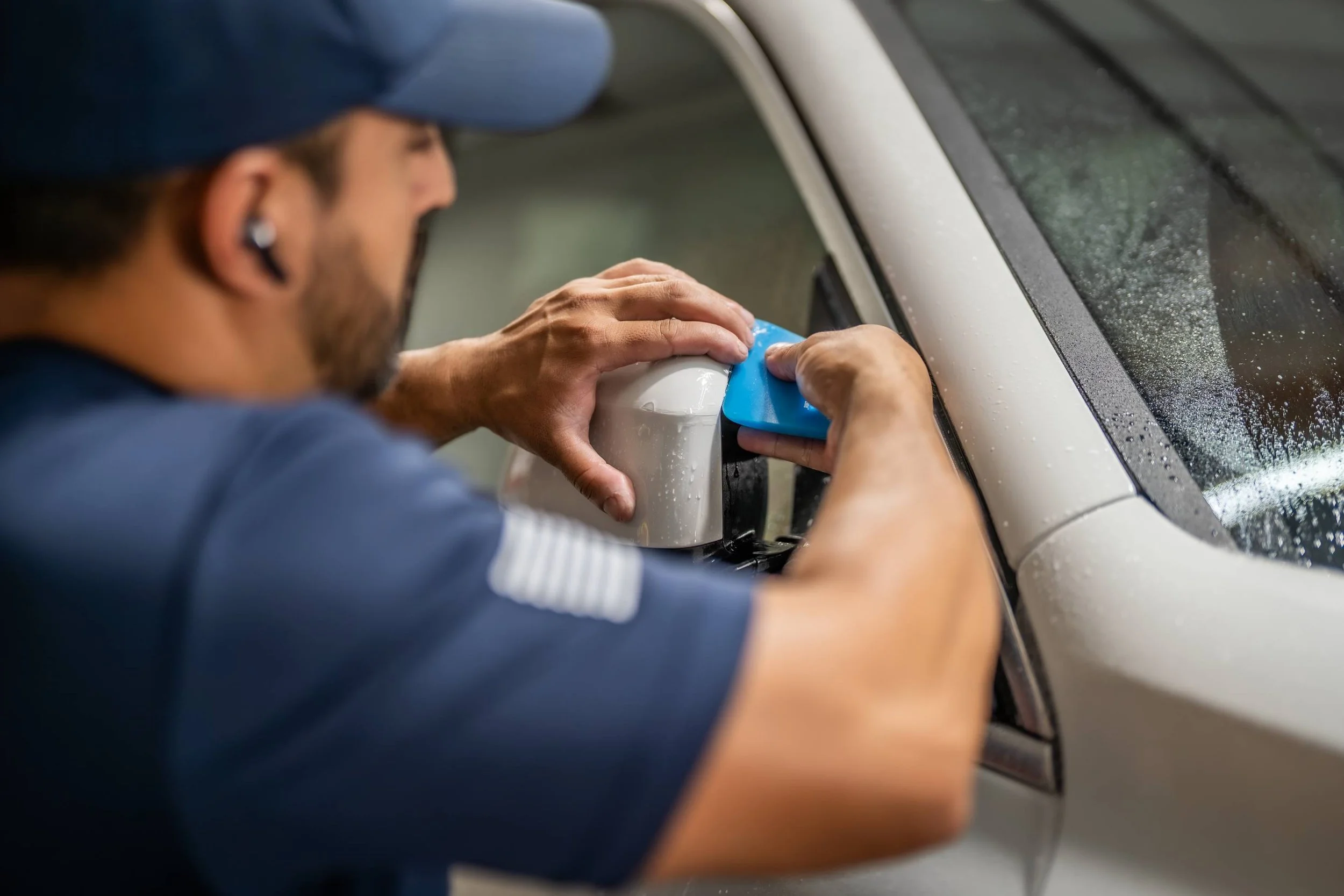 A person wearing a blue cap and blue shirt cleaning the side mirror of a white car with a blue cloth inside a garage or workshop.