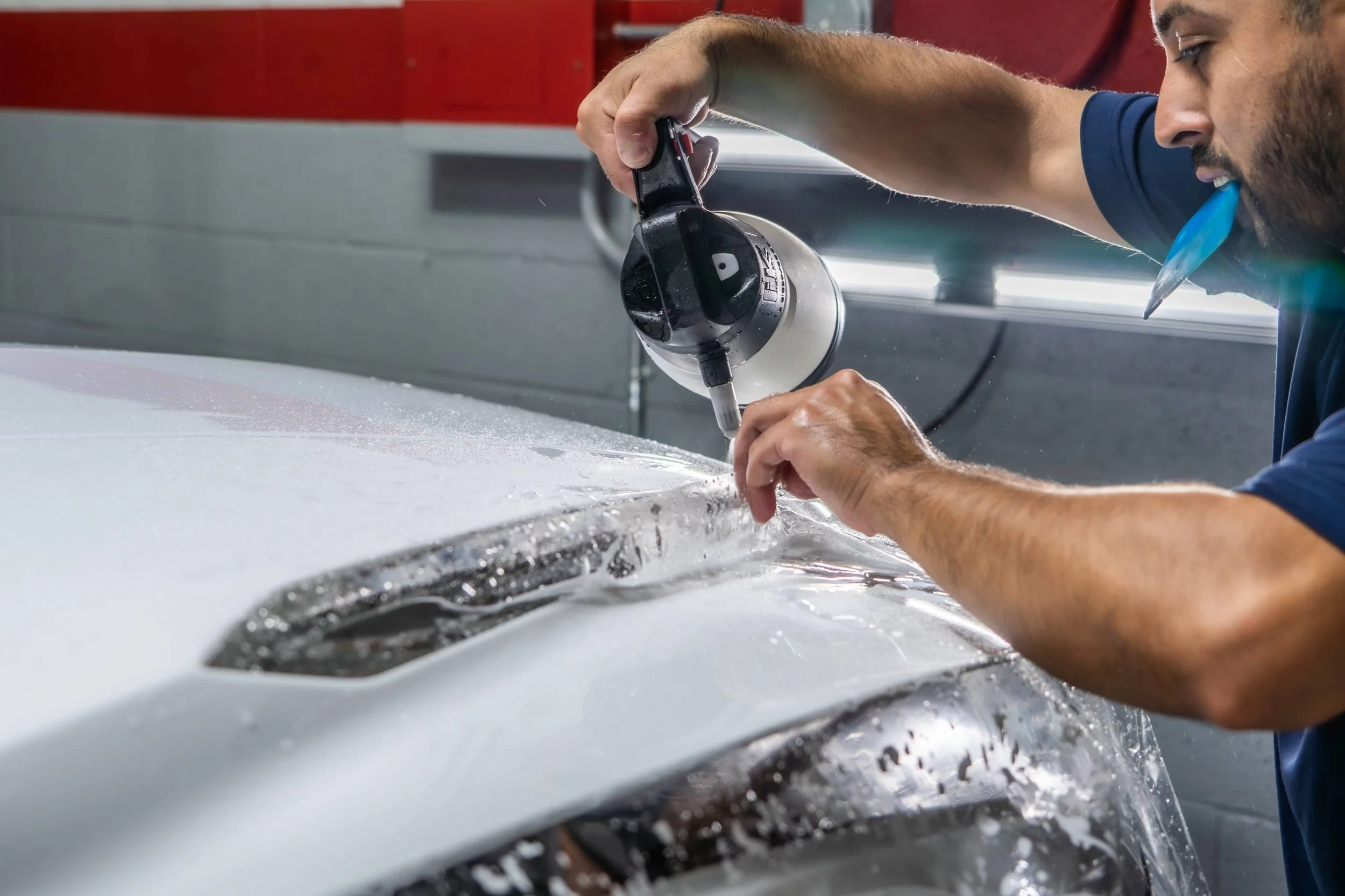 A man is using a heat gun to dry or remove paint from a white car hood.