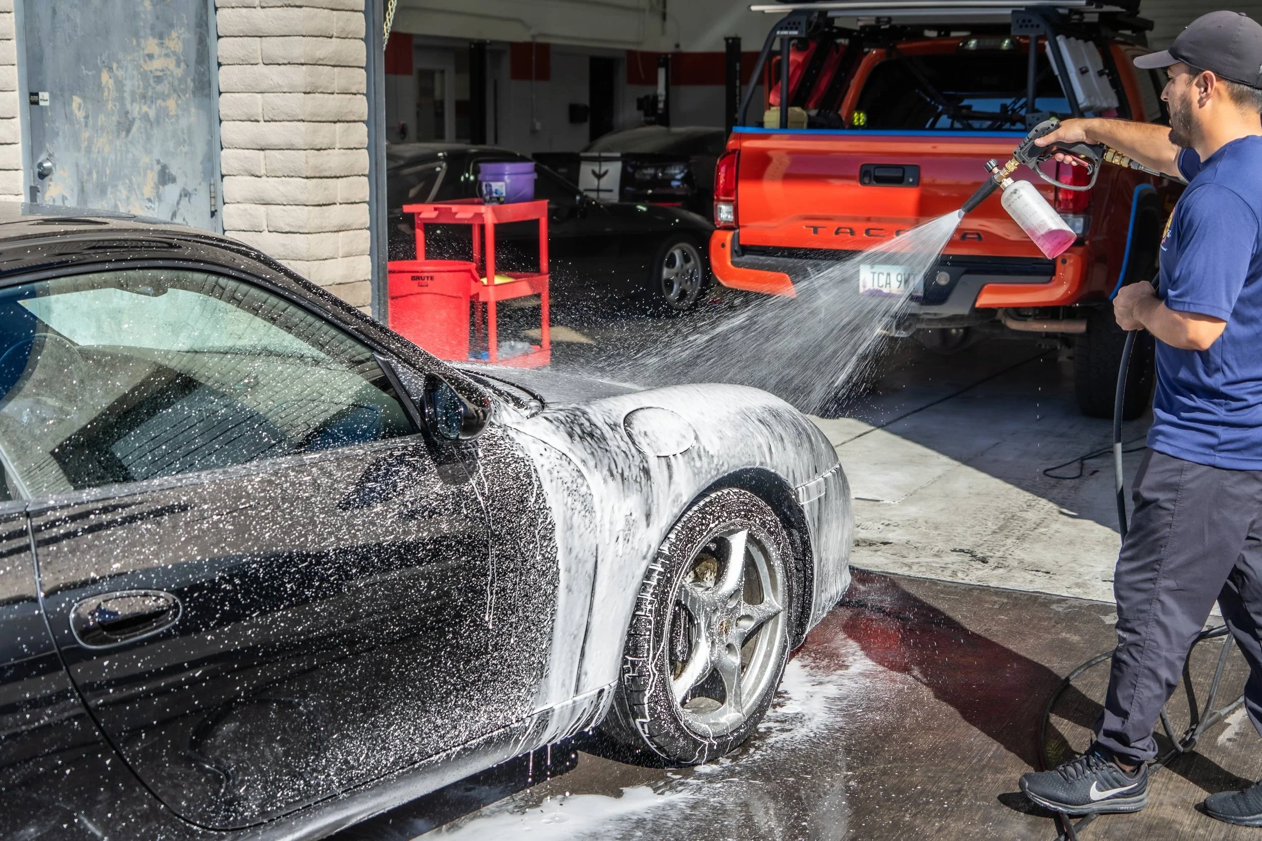 A man is washing a black sports car with soap and water at an outdoor car wash station, with a red truck and other vehicles in the background.
