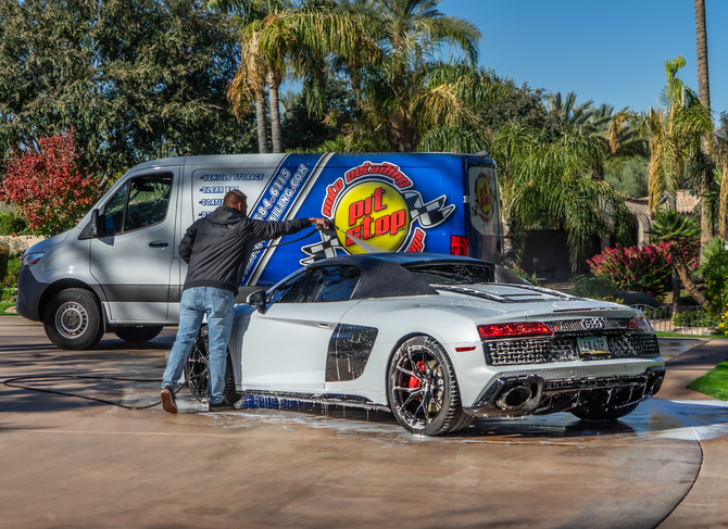 A man is washing a white Audi R8 sports car with a black roof, in front of a service van with the Pit Stop logo, on a driveway surrounded by palm trees and colorful bushes in a sunny, warm climate.