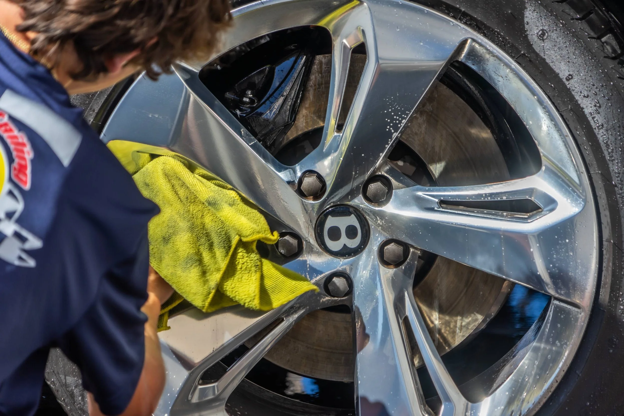 A person cleaning a chrome wheel of a luxury car with a yellow cloth.