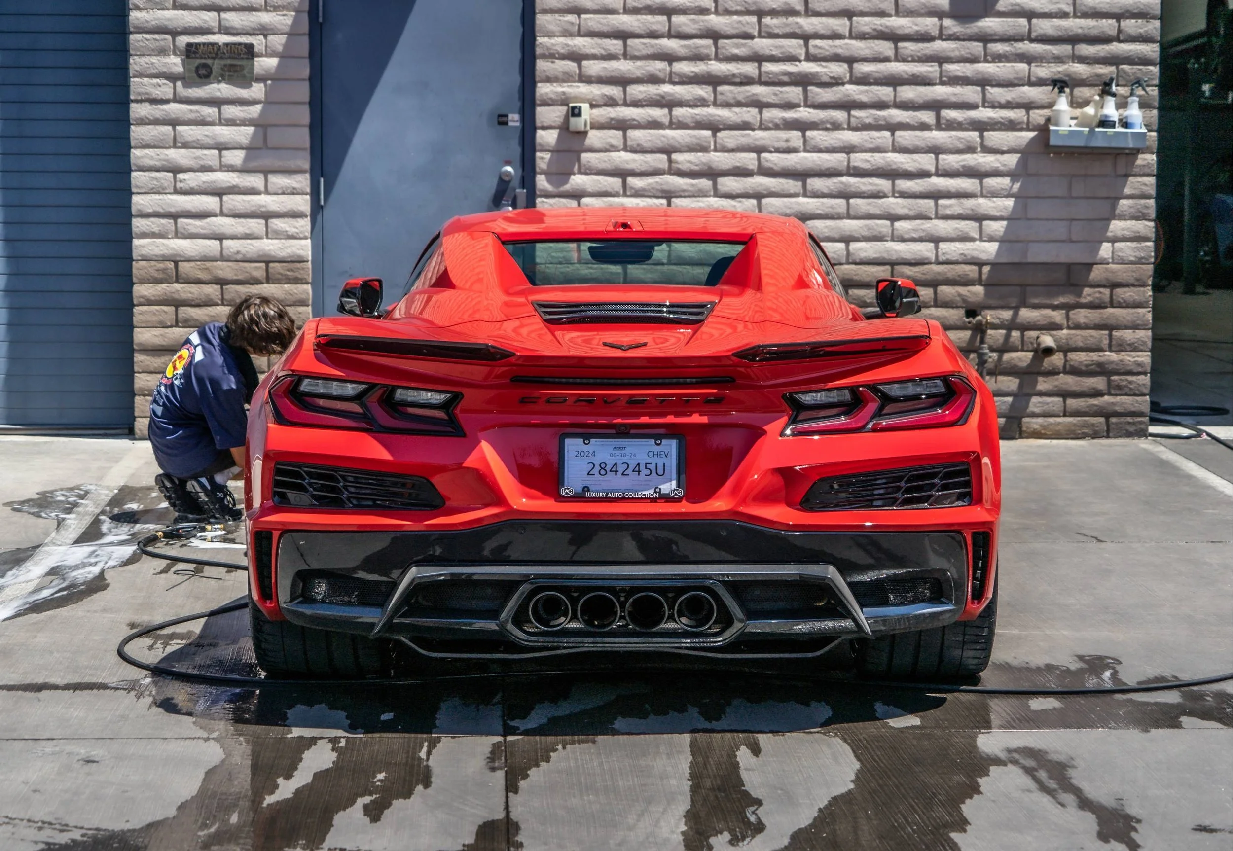 Red Chevrolet Corvette car parked outside, being washed by a person wearing a blue jacket. The car is wet with soap suds on the ground.