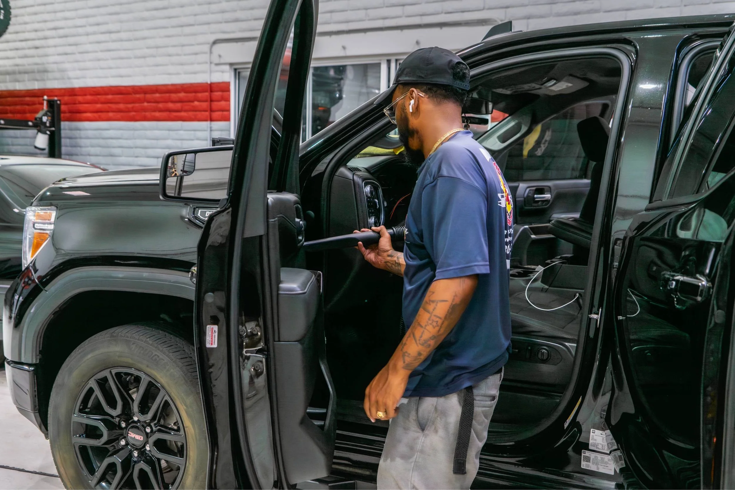 A man cleaning a black SUV with a handheld vacuum in a garage, wearing a dark cap, glasses, and a dark T-Shirt with tattoos on his arm.