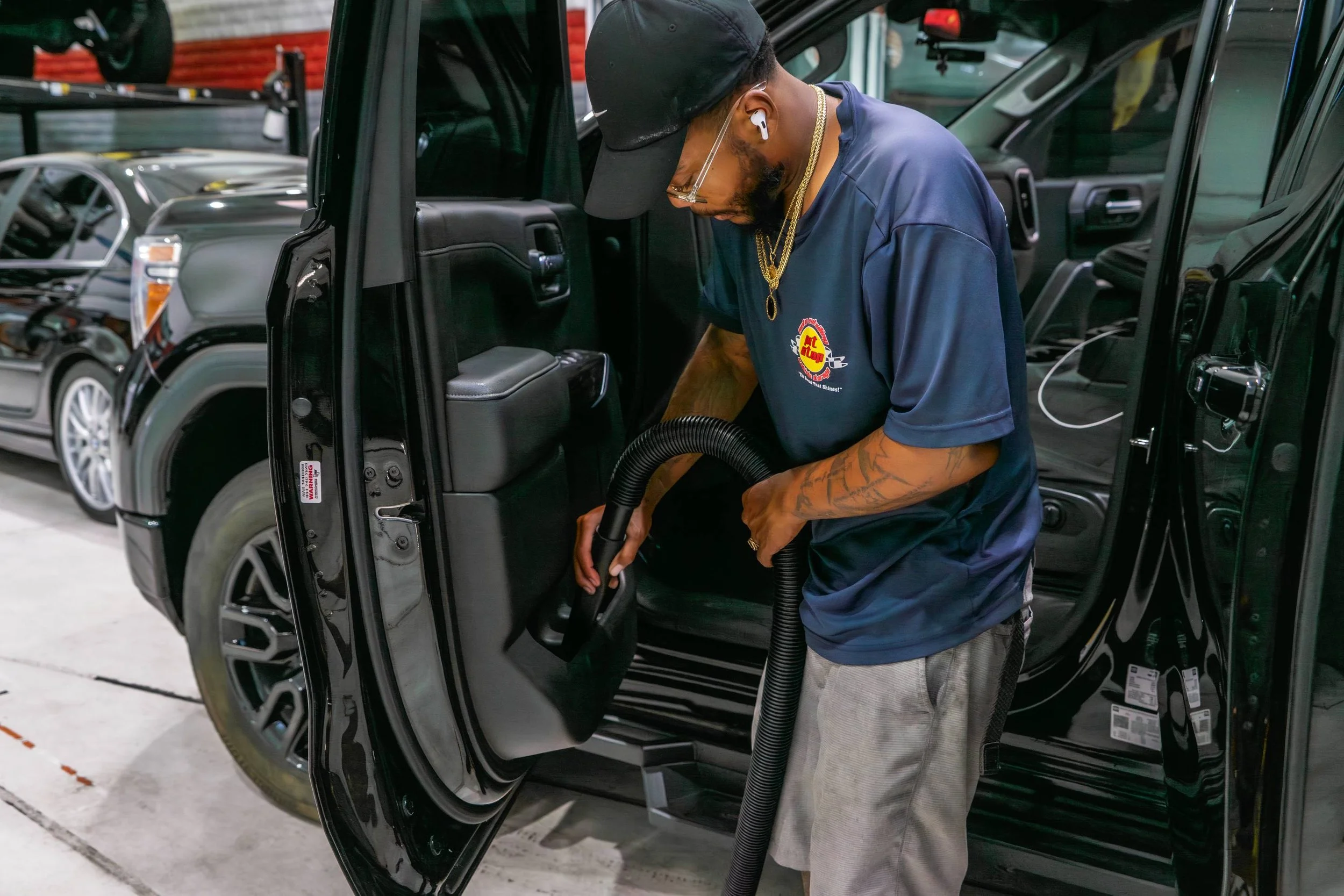 A man using a vacuum cleaner inside a black SUV in a garage or workshop with other vehicles around.