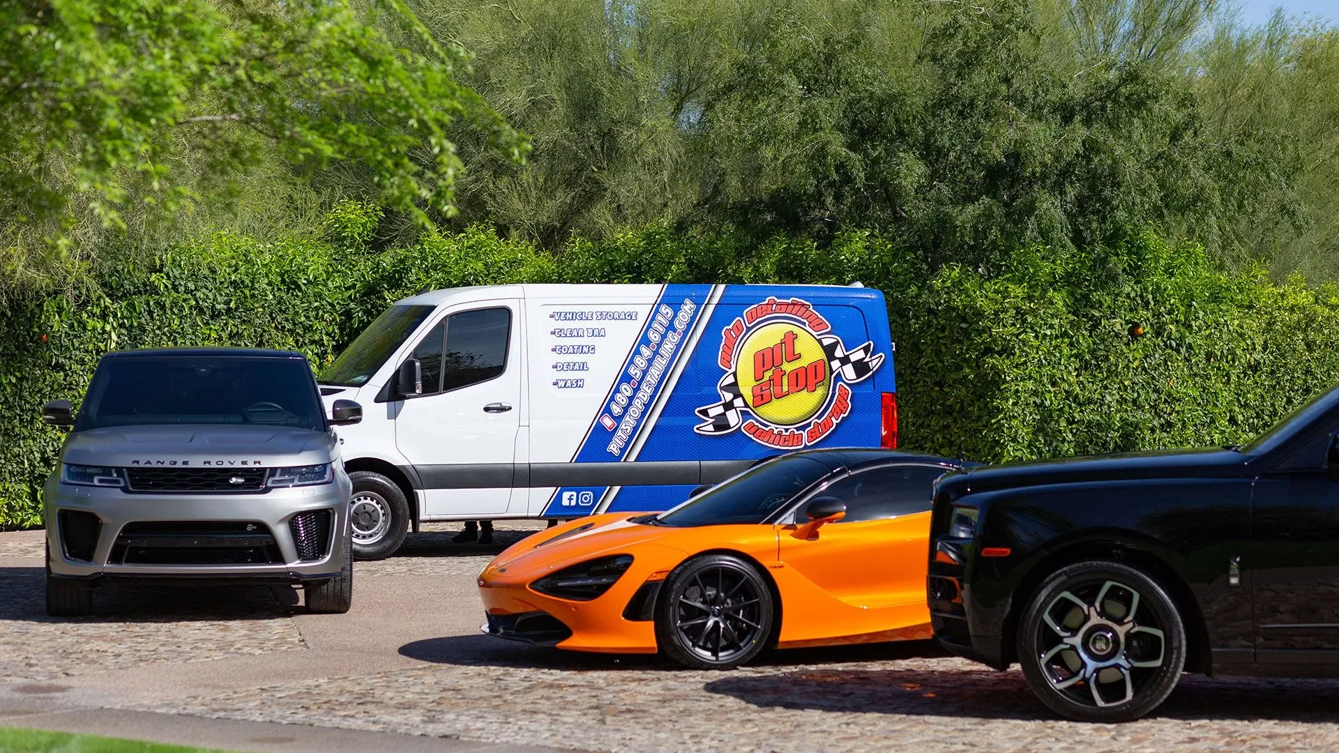 Three vehicles parked outdoors with green bushes and trees in the background: a gray Range Rover, an orange sports car, and a black vehicle. A white van with colorful graphics and advertising for a vehicle cleaning service is also in the background.