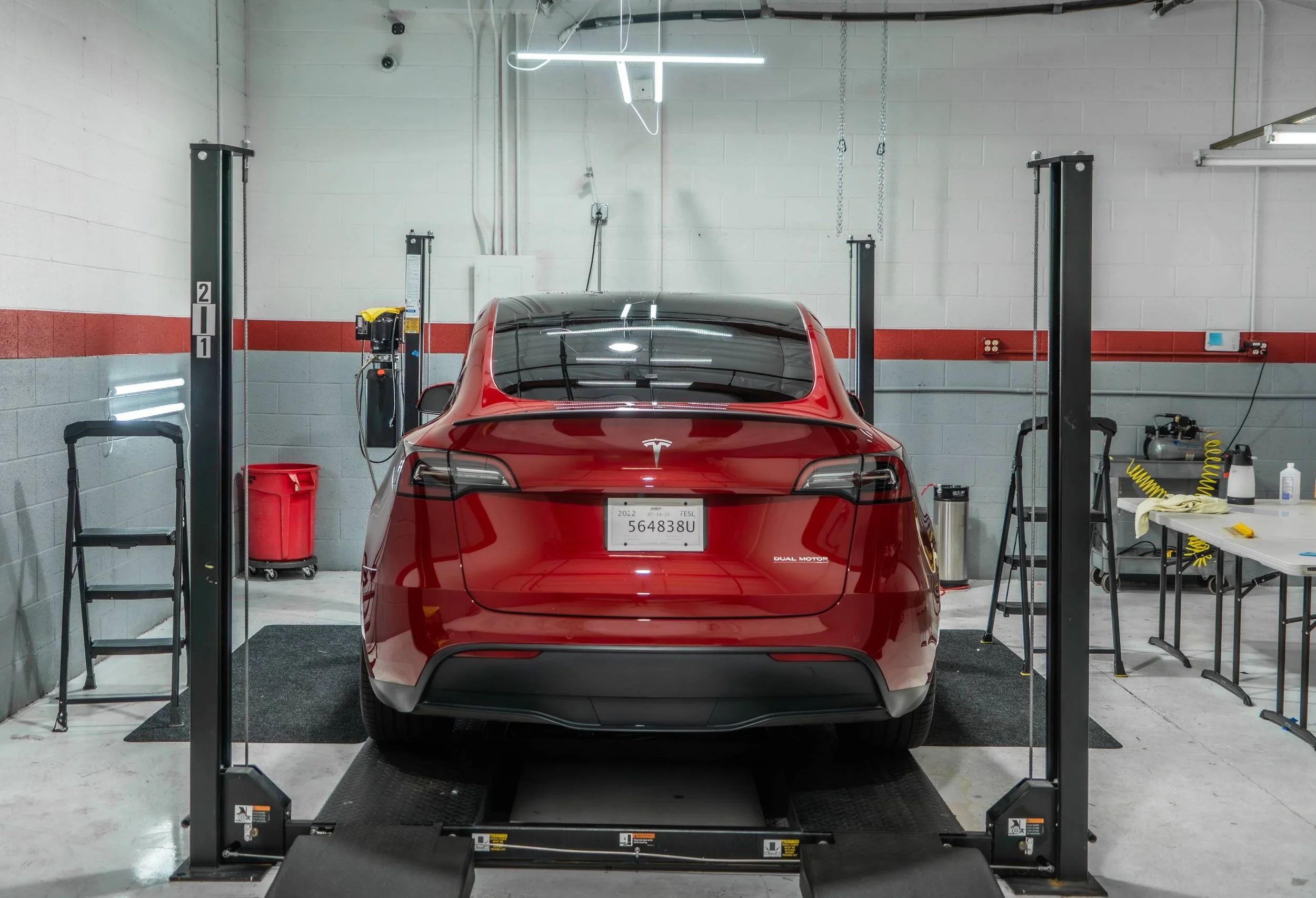 Red Tesla vehicle on a wheel alignment lift in a garage, with tools and equipment on side tables and walls, and a neon light above.