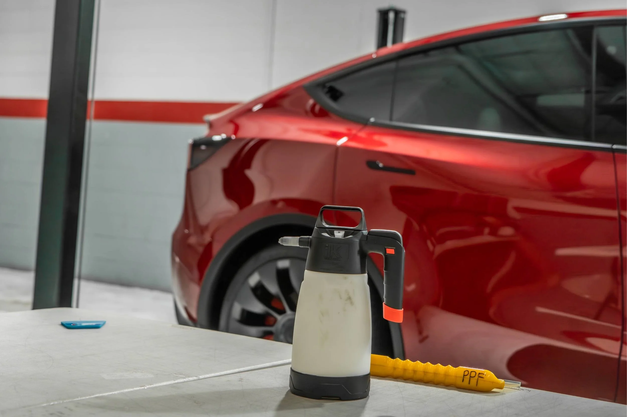 A red electric vehicle parked in a garage with a foam cannon and a yellow siphon tube on a table in the foreground.