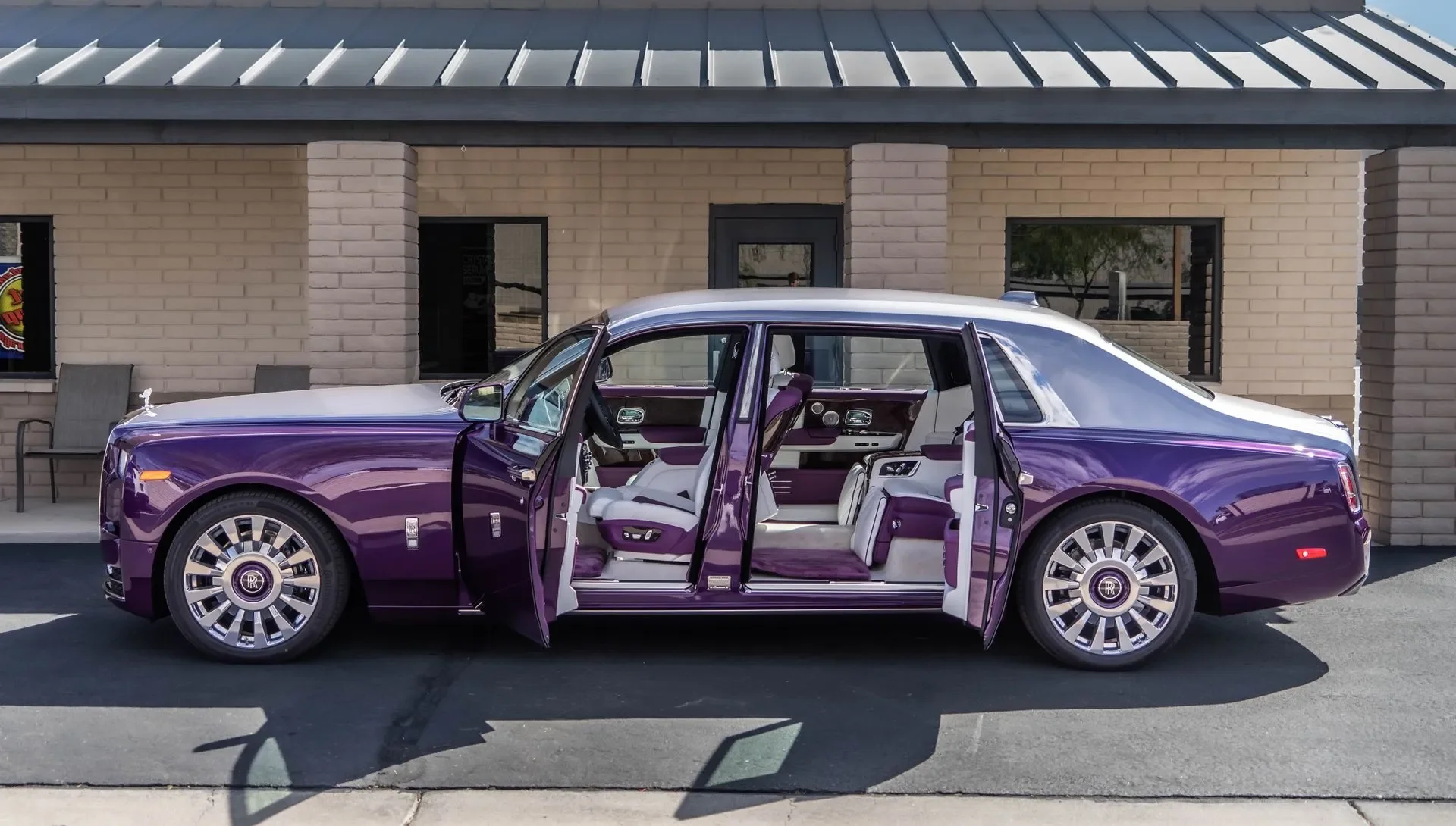 A purple luxury car with open doors showing a white and purple interior, parked outside a beige brick building with large windows and a metal roof.
