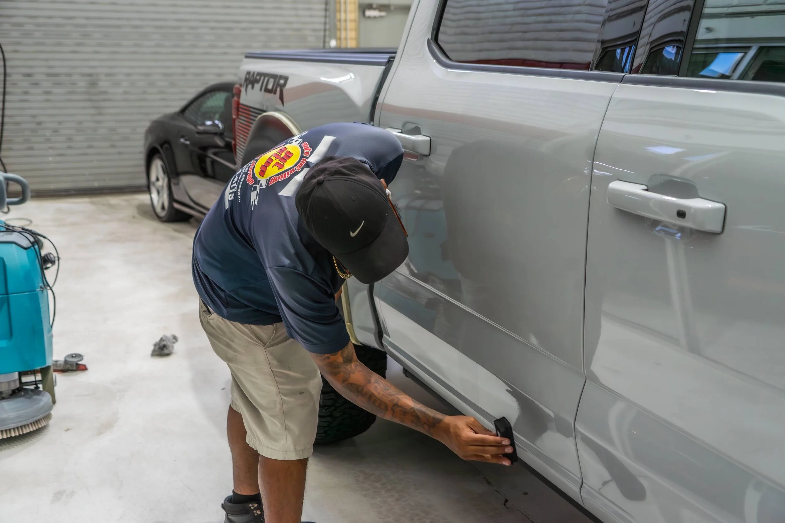 A person wearing a black cap, blue Nike hoodie, and shorts is working on the side of a silver car in a garage. They are crouched down, focused on the task, possibly repairing or inspecting the vehicle. There is a black car and a closed garage door in the background.