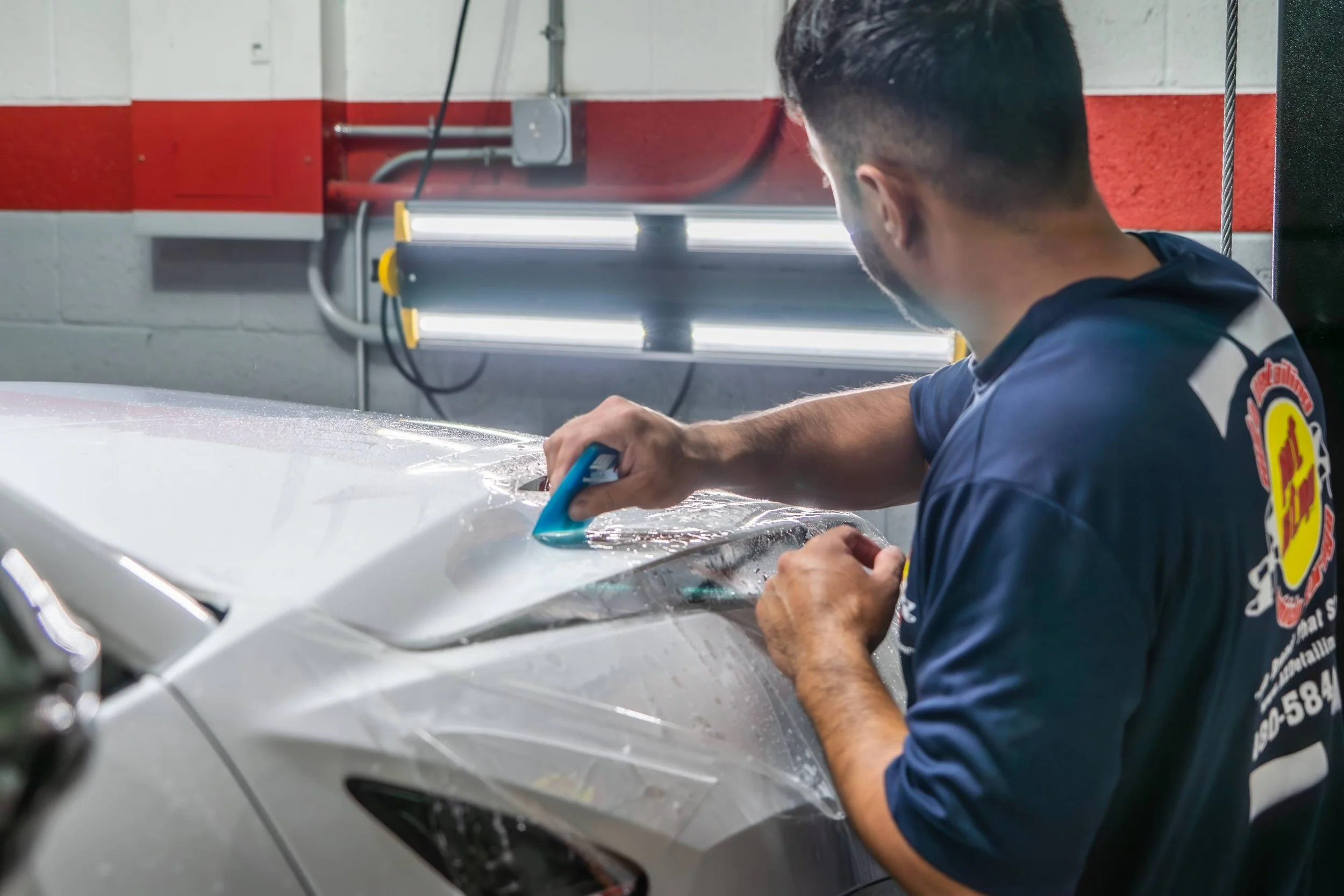 A man working on a car, using a tool on the hood, in an indoor garage with fluorescent lights and red and white painted walls.