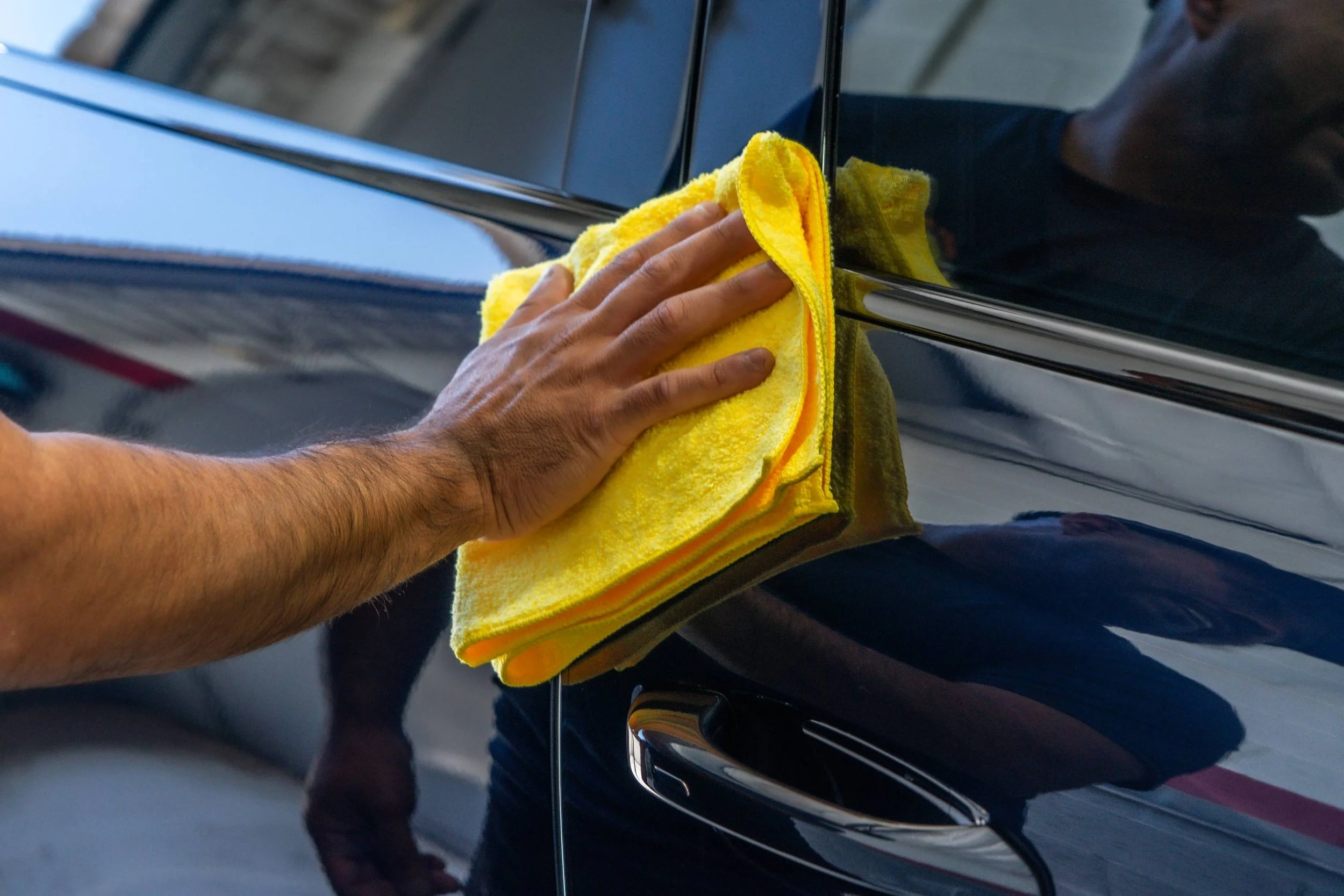 Person cleaning a black car door with a yellow microfiber towel.