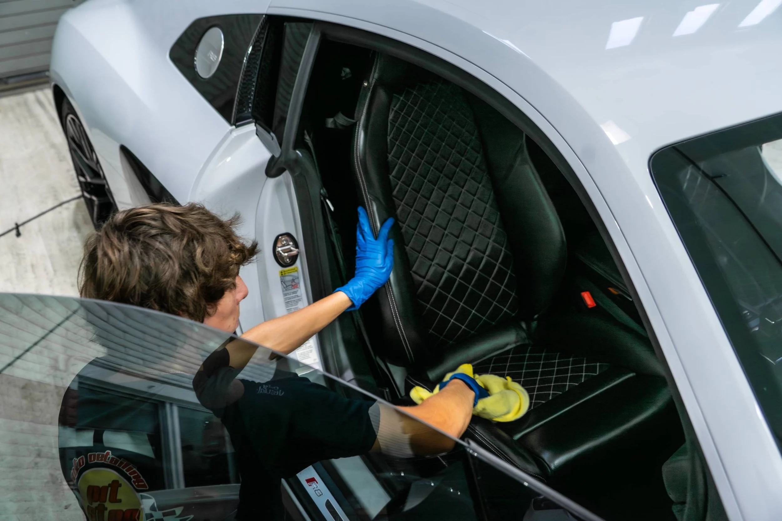 A person wearing a black shirt and blue gloves cleaning the interior of a silver car, focusing on the seat.