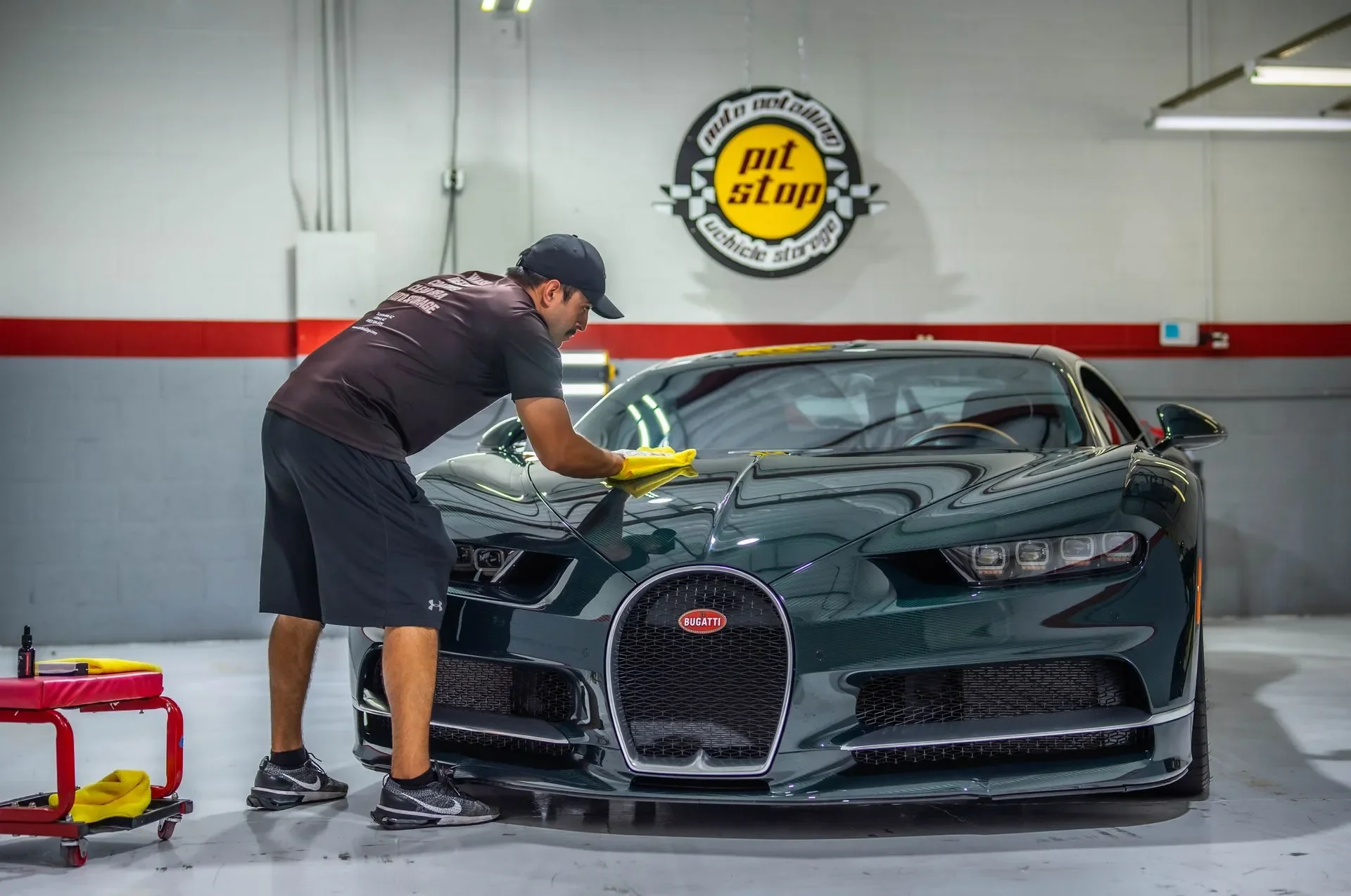 A man cleaning a black Bugatti sports car in a garage with a sign that reads 'pit stop' on the wall.
