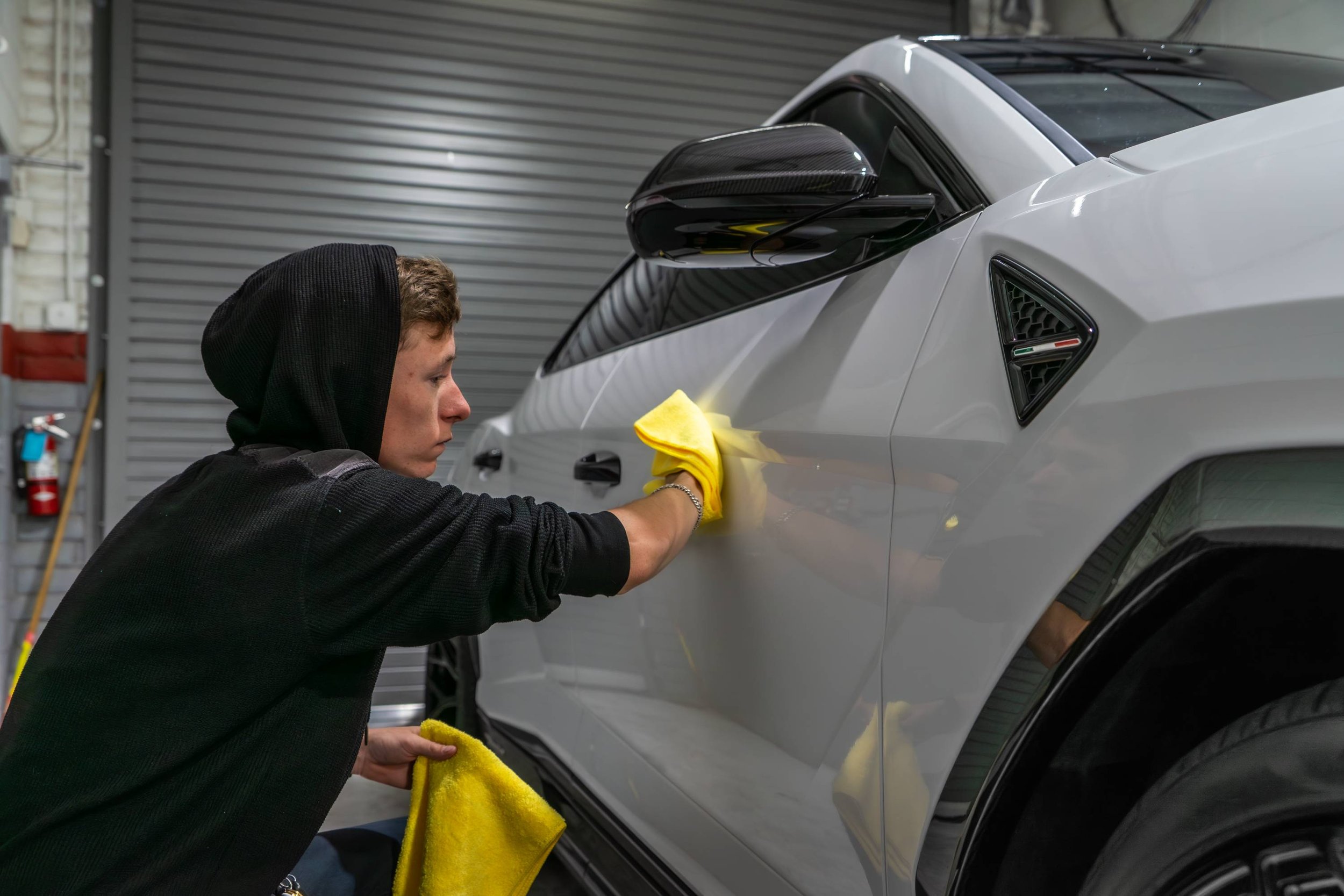 A person cleaning a grey car with a yellow cloth in a garage.