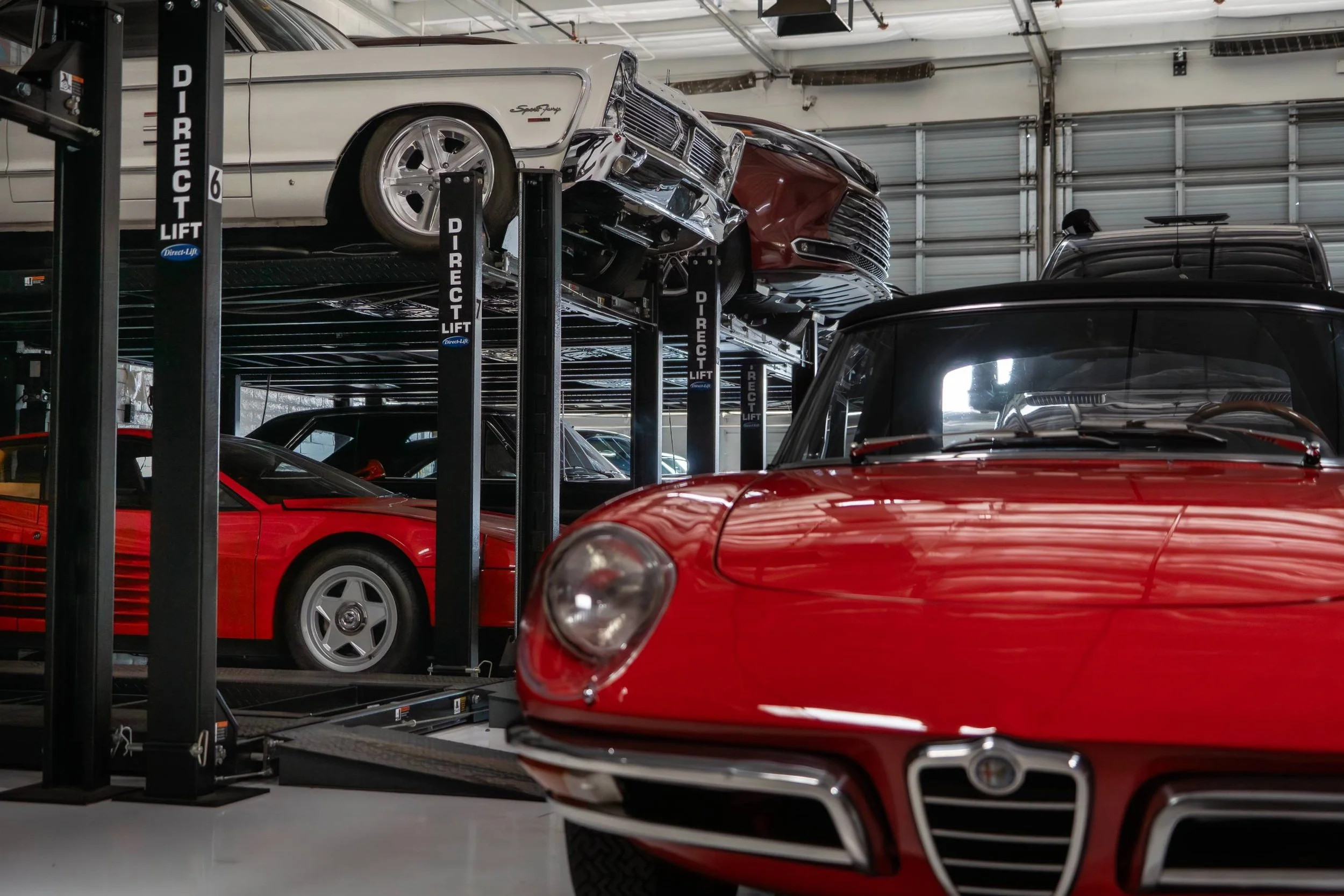 An indoor car storage facility with multiple cars on hydraulic lifts. A white vintage car is on the upper lift, and a red modern sports car is on the lower level. The garage has high ceilings and large overhead doors.
