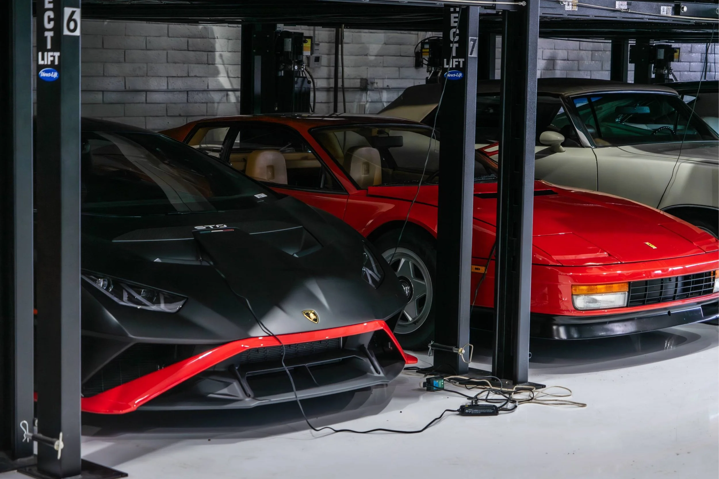 A storage unit with luxury cars, including a black Lamborghini and a red Ferrari, parked inside. The cars are on metal shelving, and there are various electrical cords and equipment on the floor.