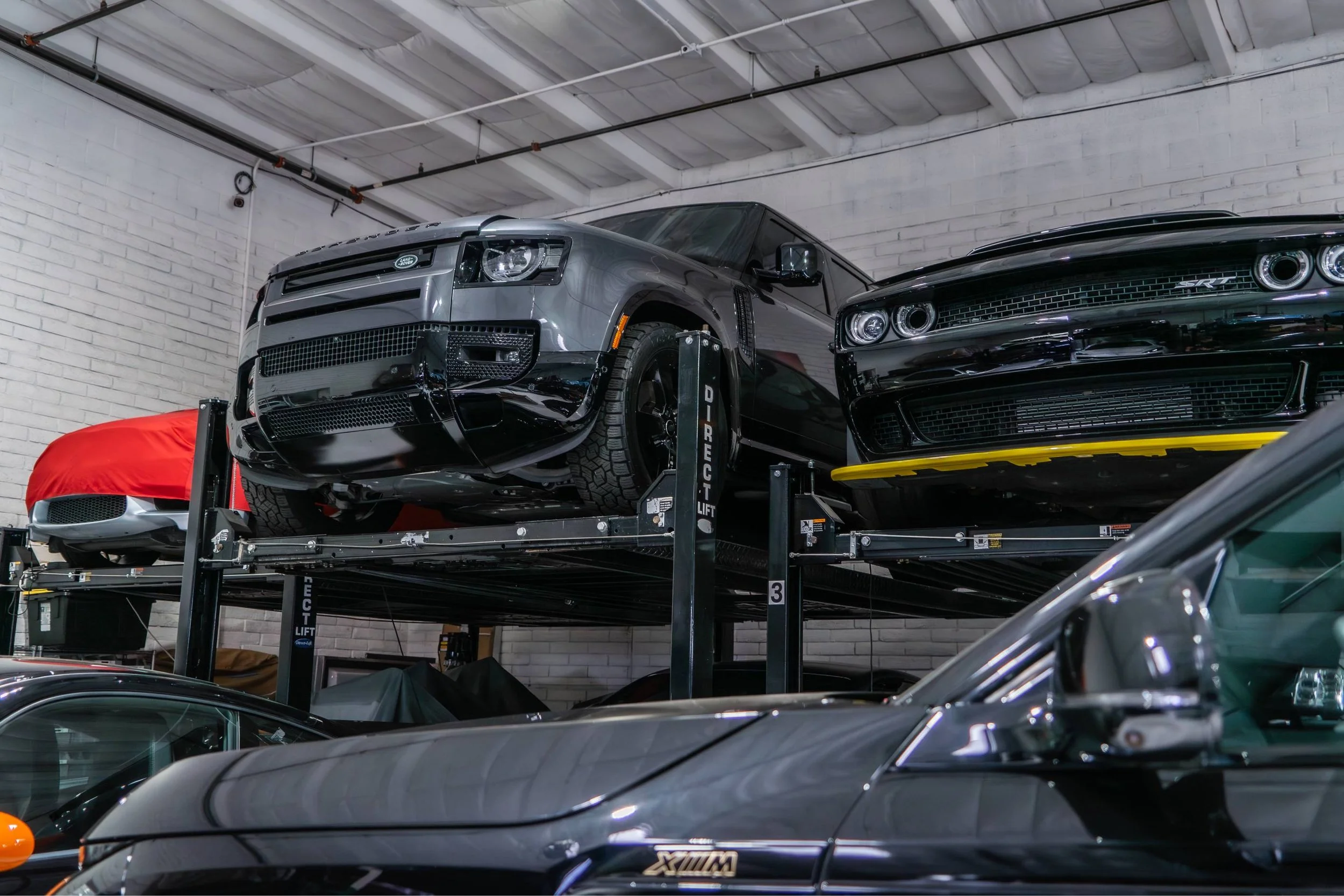 Two cars, a black Dodge Charger and a gray Range Rover, parked on a car lift inside a garage with white brick walls and a metal ceiling.