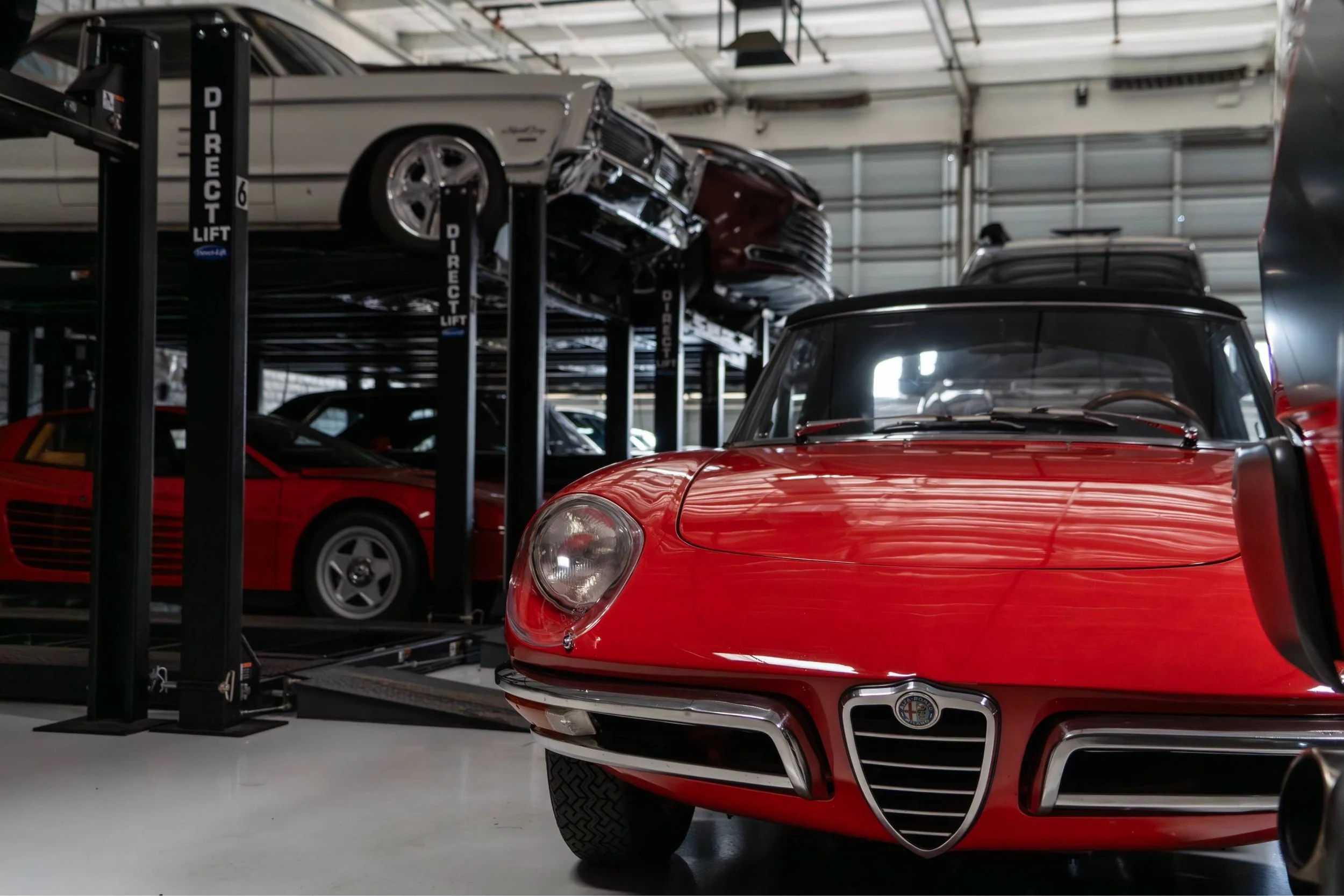 A red classic car, an Alfa Romeo, in the foreground inside a garage. Behind it, a black car is on a hydraulic lift, with another classic car, possibly a vintage model, on top of the lift.