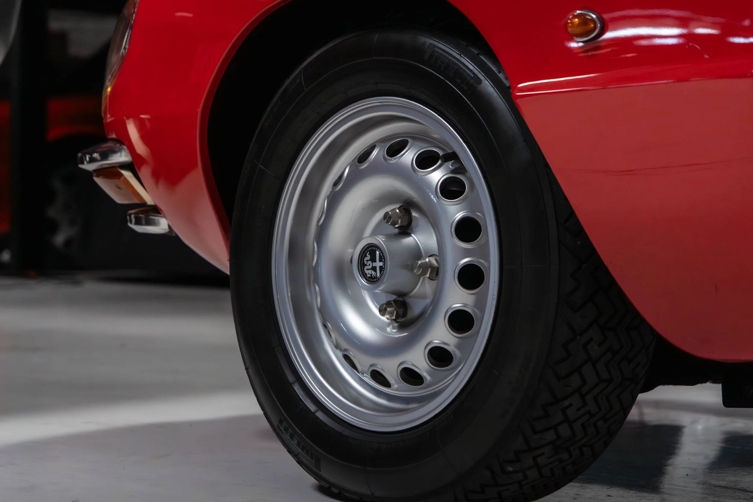 Close-up of a vintage red car's front wheel with a silver steel rim and black tire, showing part of the car's red body and bumper.