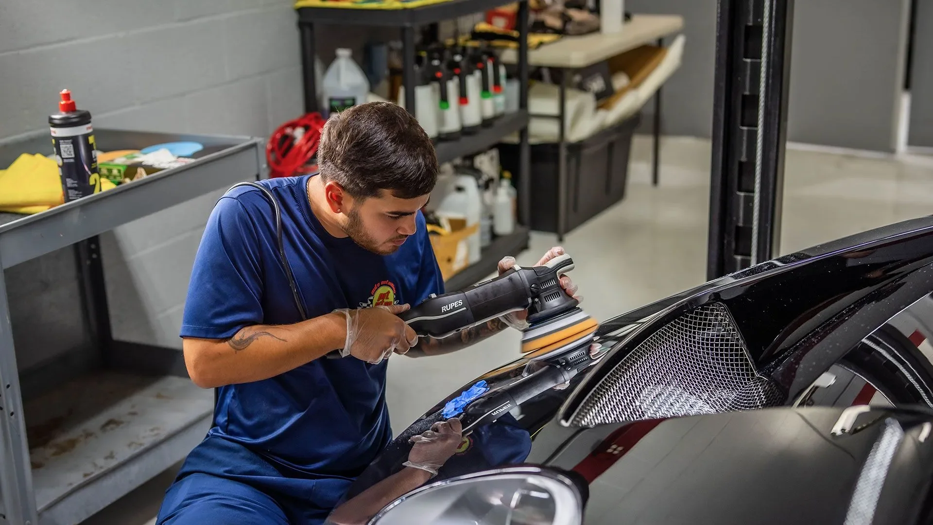 A man polishing a black sports car in a garage or workshop with various tools and supplies on shelves in the background.