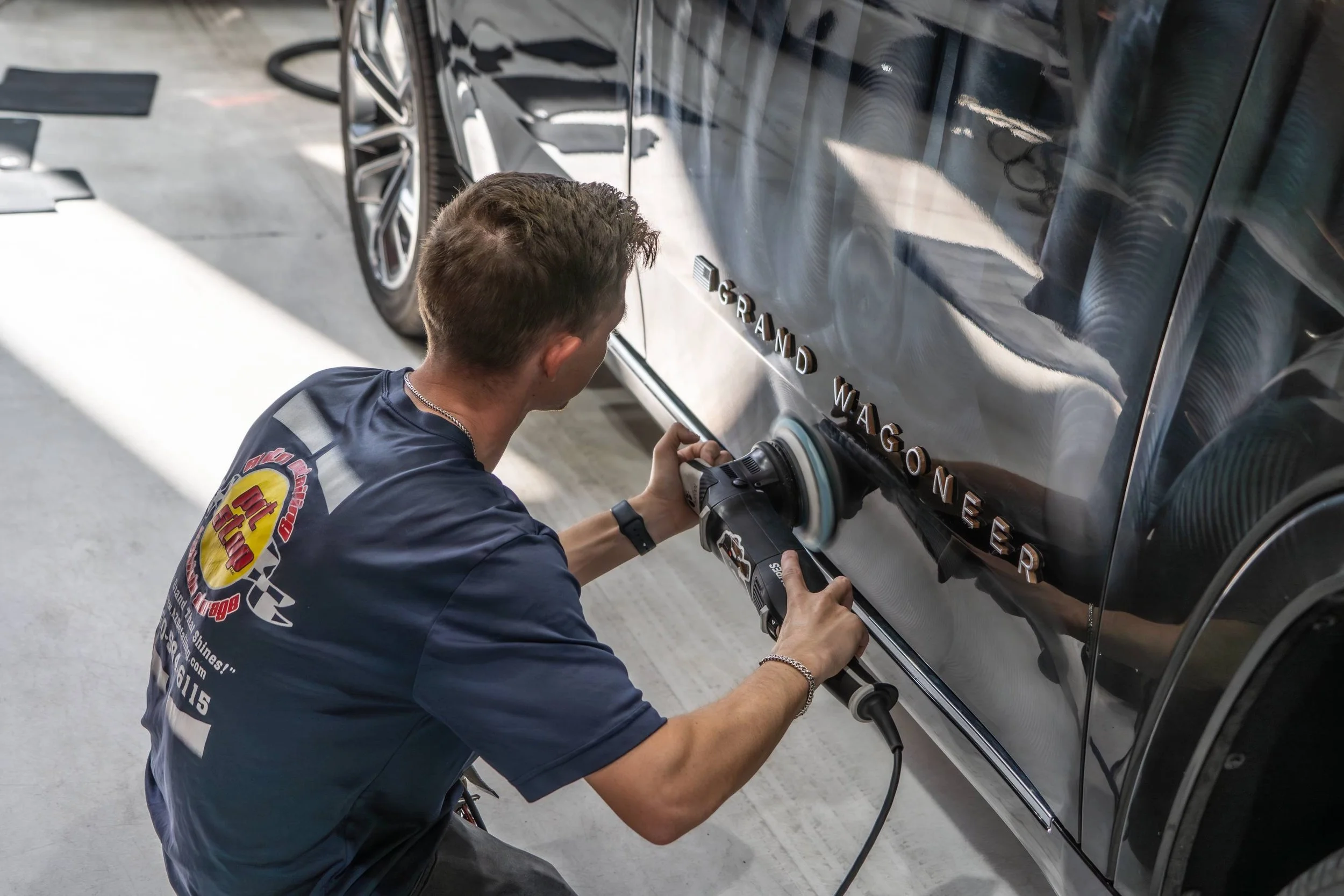 A man polishing the side of a black vehicle with a power buffer, spelling out 'Grand Wagoner' in chrome letters.