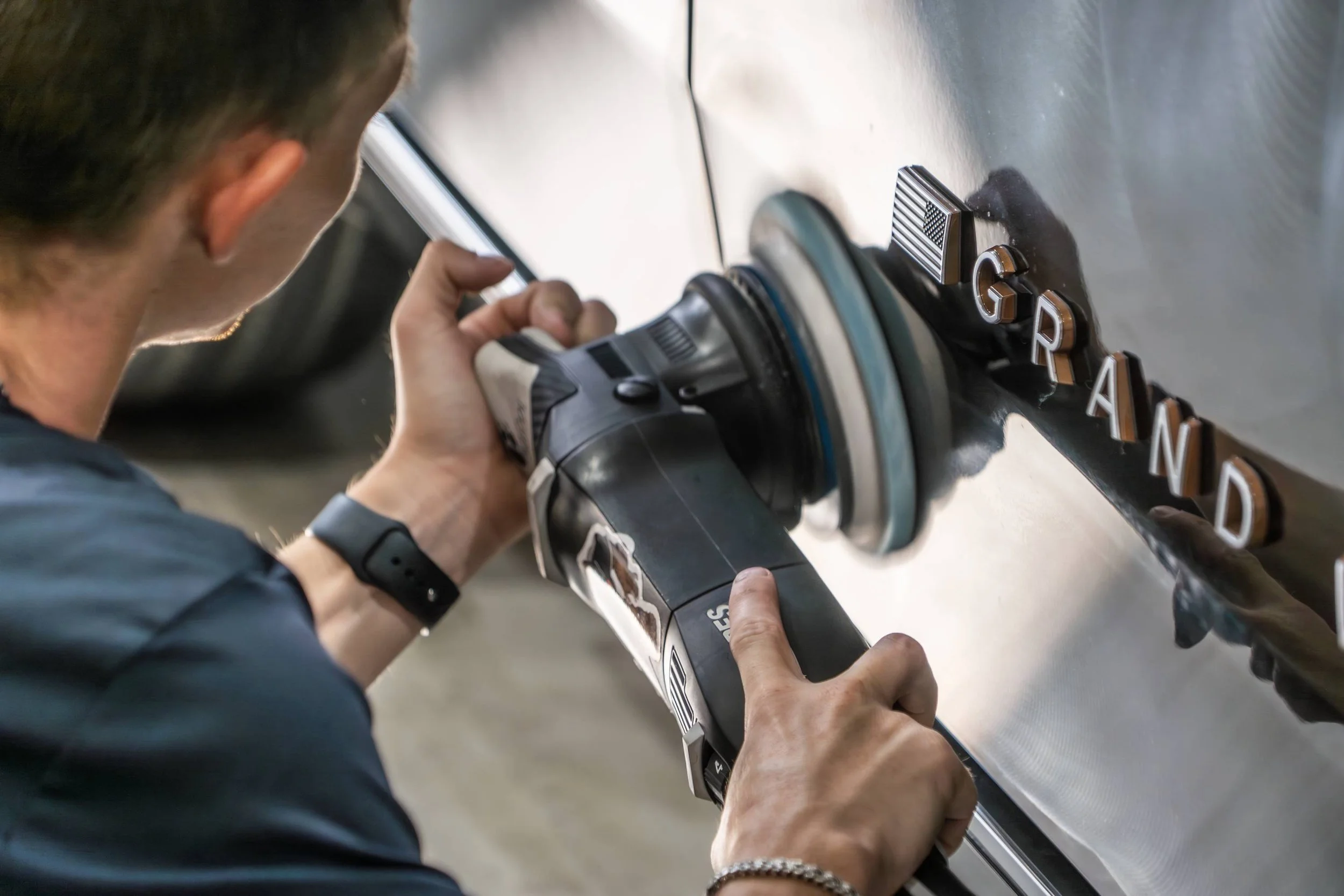 A person polishing a metallic surface with a handheld buffer, spelling out 'GRAND' with individual letters on the surface.