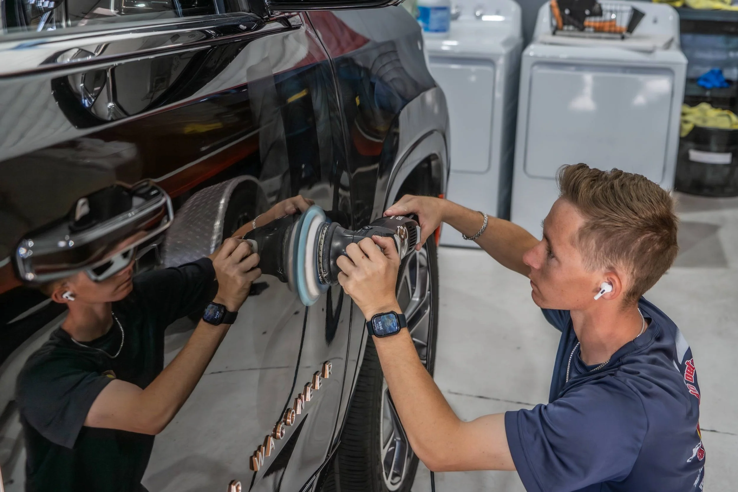 Two workers polishing a black car with a power buffer in a garage or workshop.