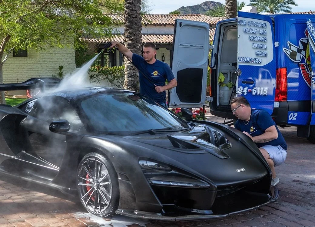 Two men cleaning a black sports car with a pressure washer, with a cleaning truck nearby.